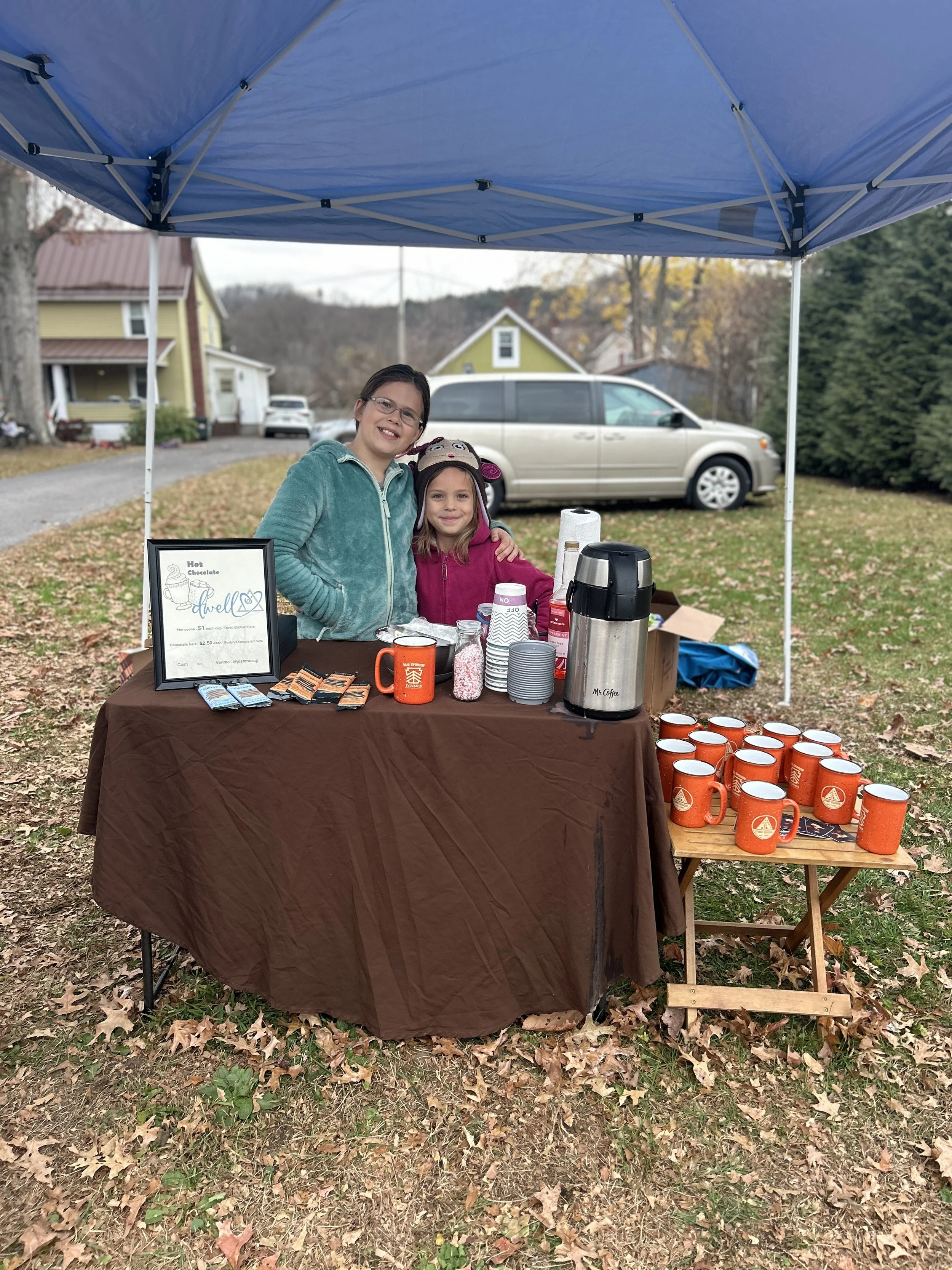 Two young girls standing behind a hot chocolate stand outdoors under a blue canopy, with a brown tablecloth. The stand has coffee mugs, packets of hot chocolate, cups, and a large coffee urn. The girls are smiling, one wearing a teal jacket and glass