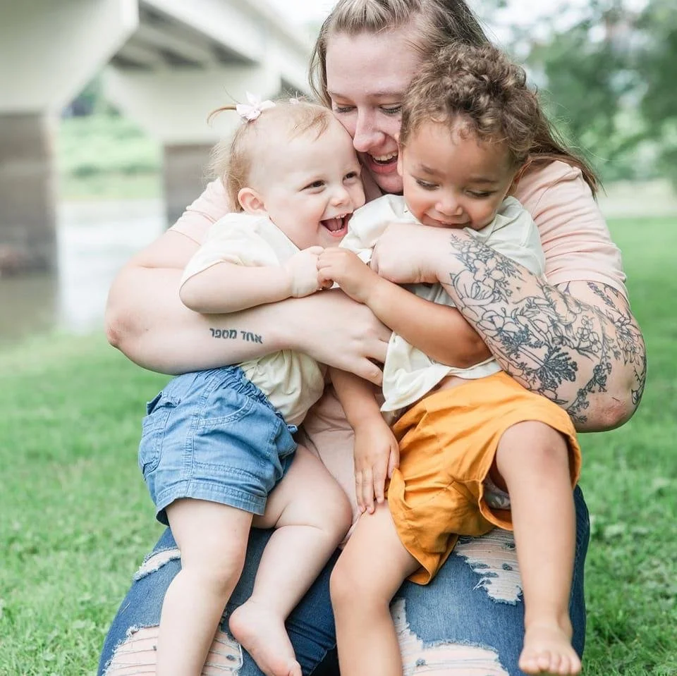 Woman sitting on grass hugging two smiling children of different races.