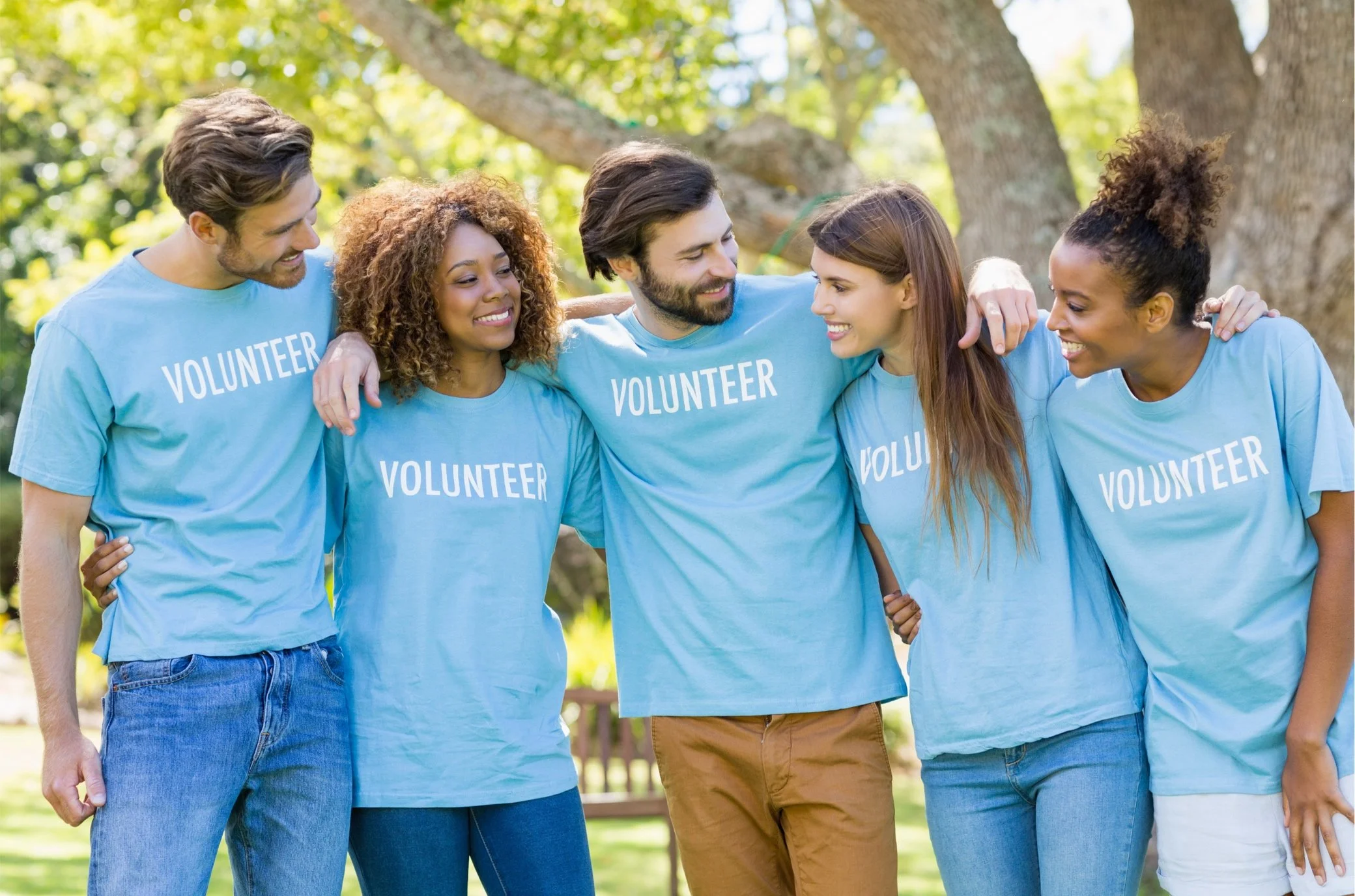 Group of six diverse volunteers standing outside, wearing matching light blue t-shirts with the word 'VOLUNTEER' printed on them, hugging and smiling at each other.
