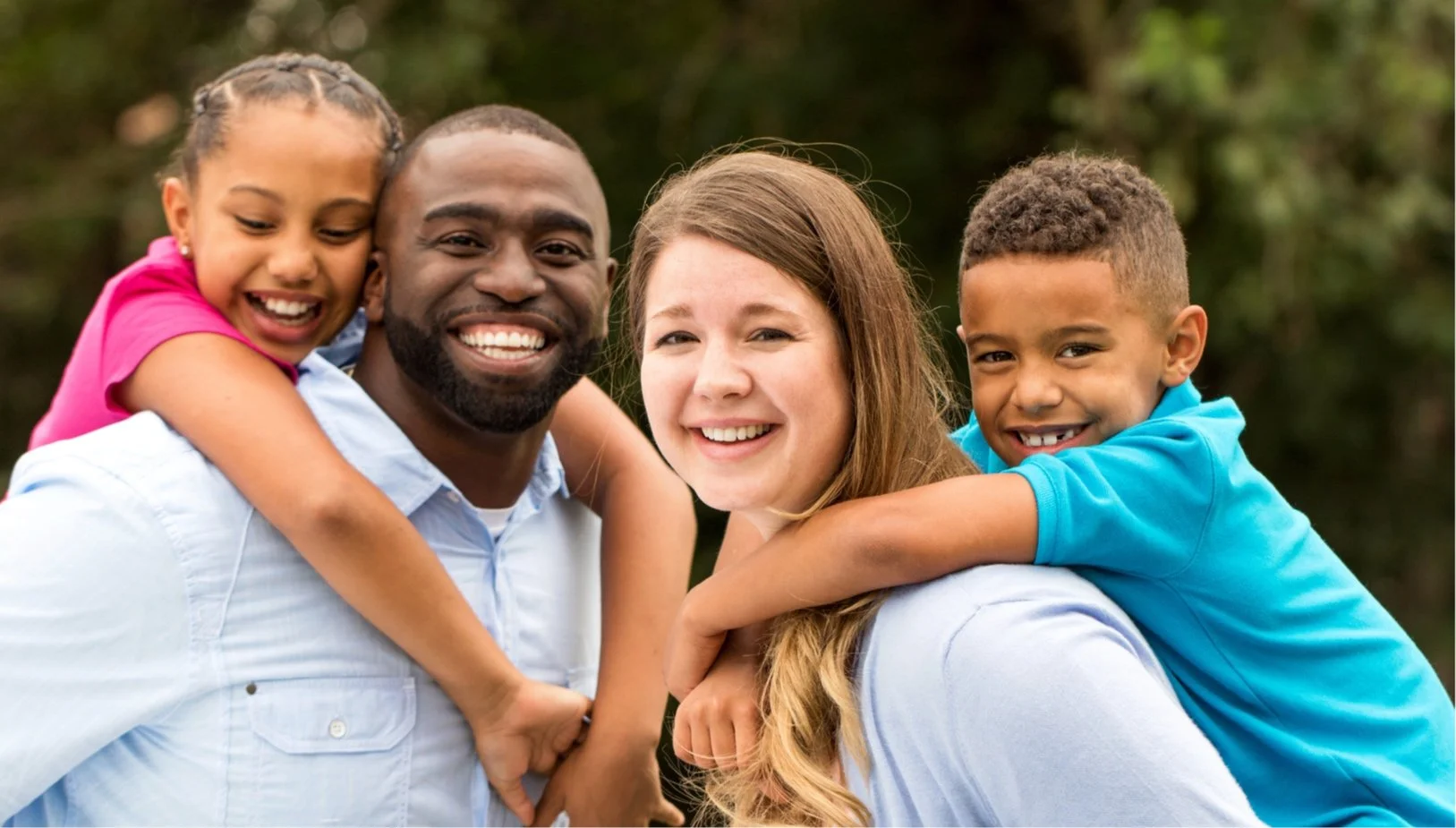 A happy family of four outdoors, with children riding on their parents' shoulders, smiling.