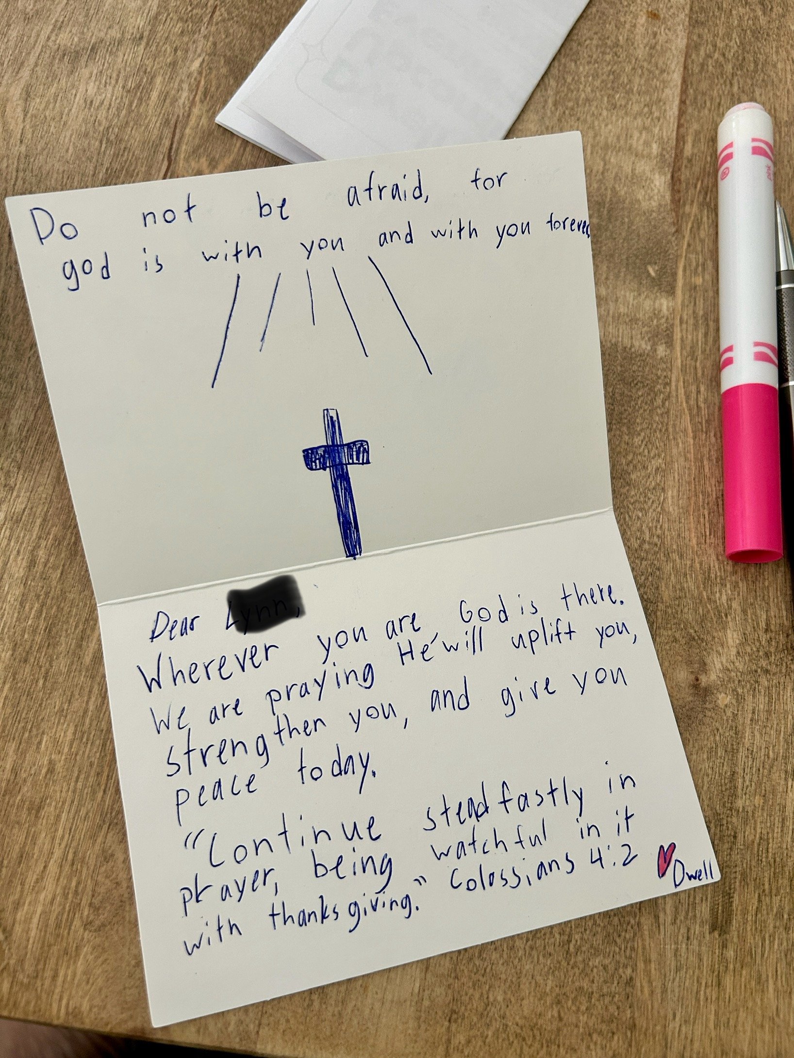 A handwritten note on a white card with a drawing of a cross and a prayer or message, placed on a wooden surface. A pink marker and another pen are nearby.