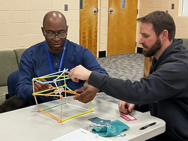 Two men are playing with a colorful stick and string structure model at a table in a room with beige brick walls and two closed doors in the background.