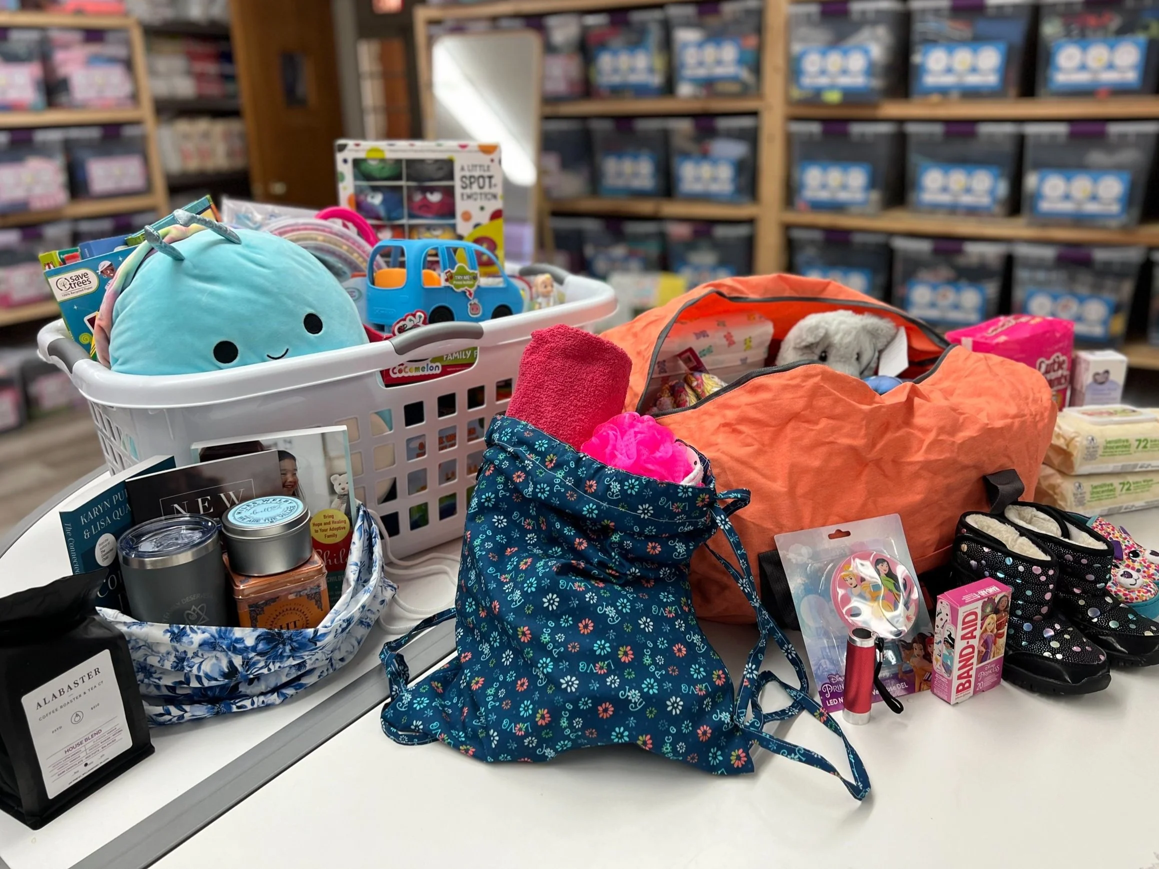 A table with baskets and bags filled with various items including toys, games, children's books, essential care items, clothing, shoes and caregiver support items. The background shows shelves with storage containers.