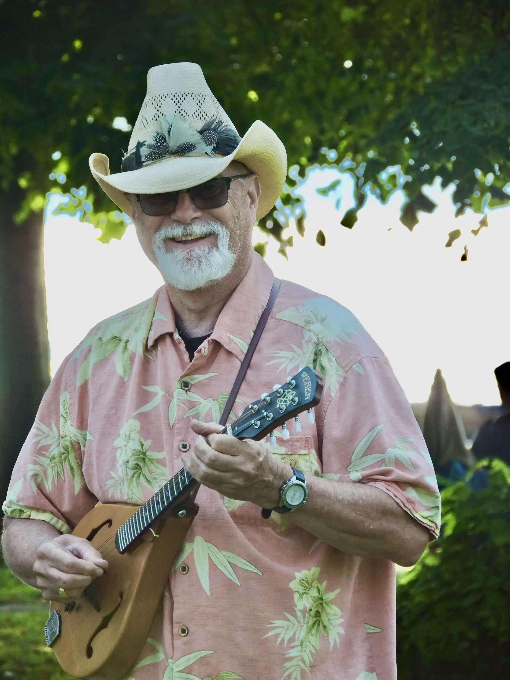 A smiling man wearing sunglasses and a cowboy hat decorated with feathers, playing a small stringed instrument outdoors near a tree.
