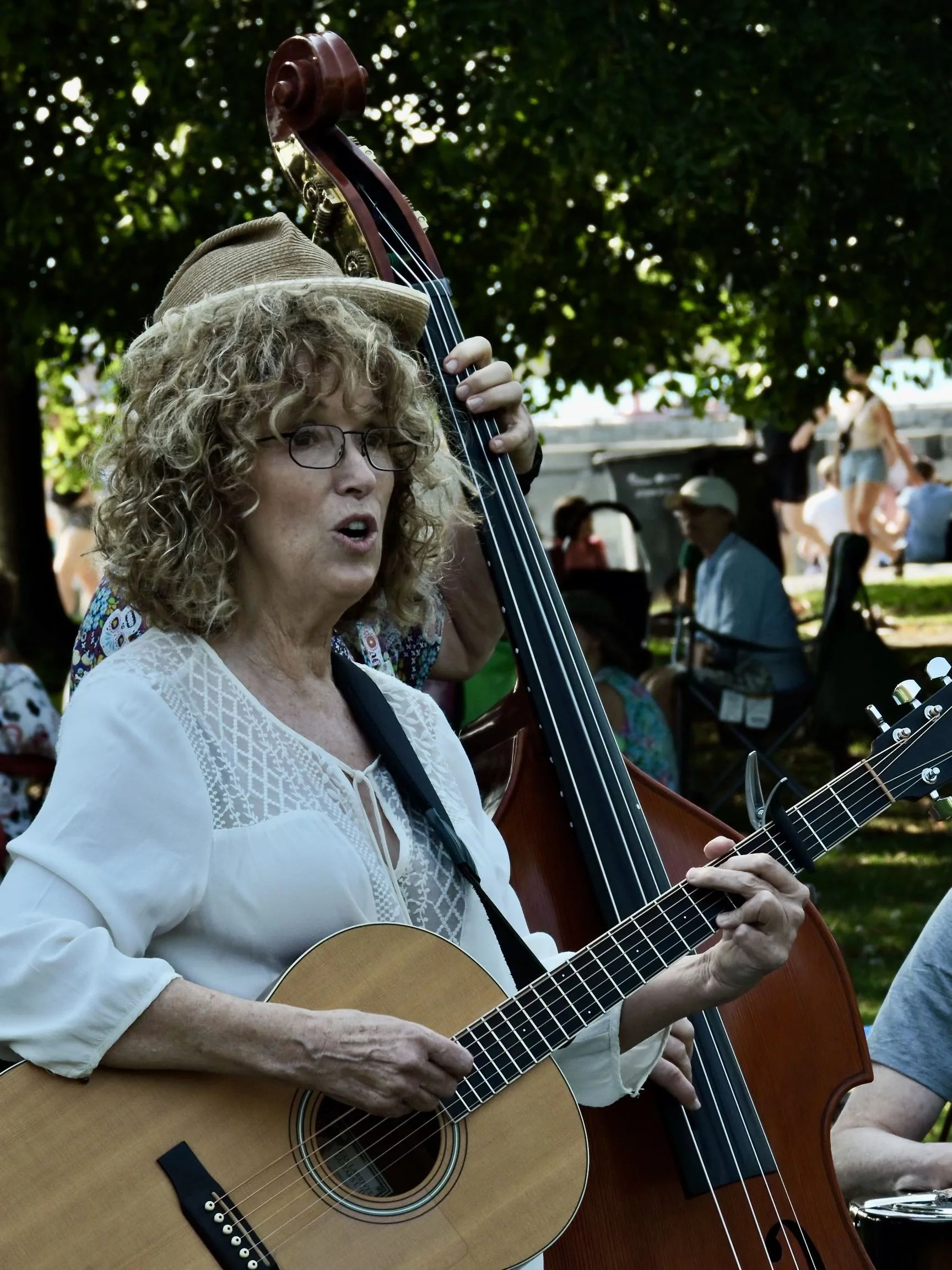 A woman with curly blonde hair, glasses, and a beige hat plays an acoustic guitar and sings outdoors during a gathering or festival, with a double bass behind her and people sitting and walking in the background under trees.