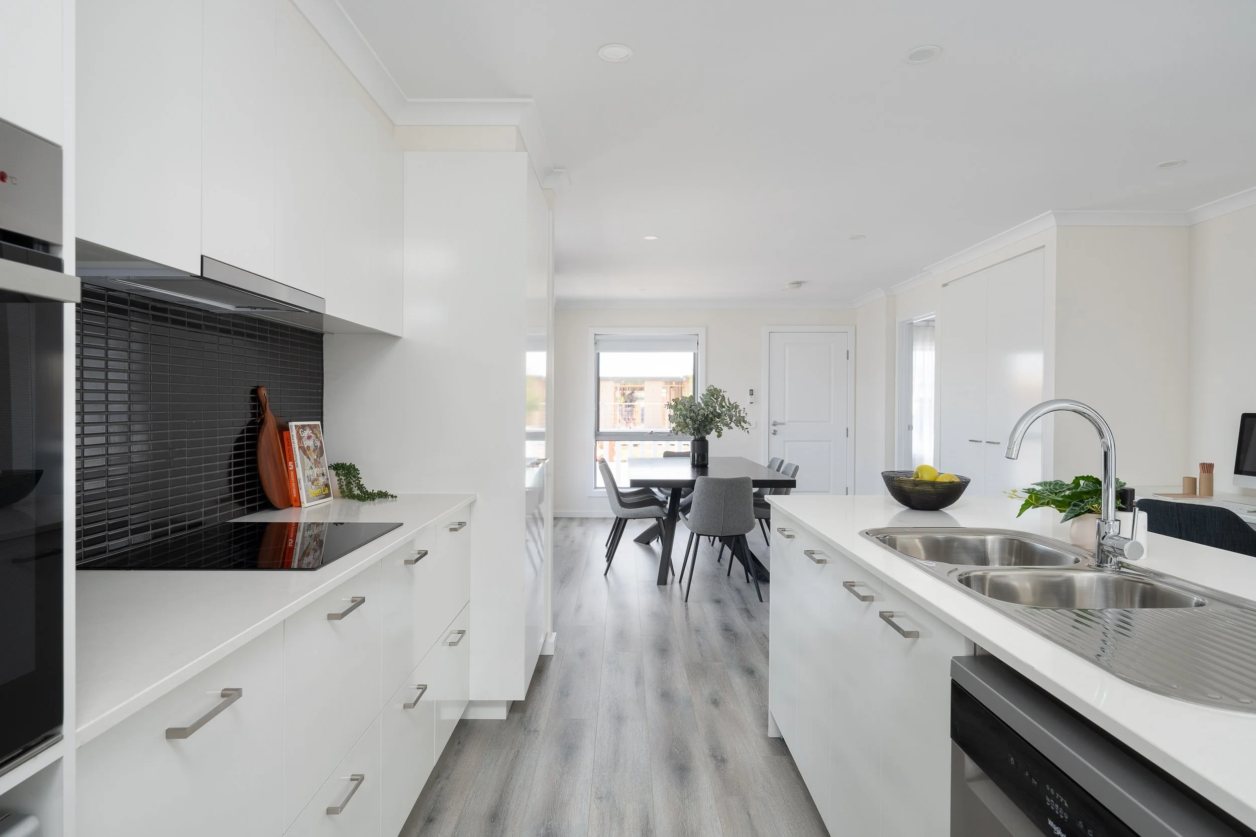 Modern kitchen interior with white cabinetry, black backsplash, and a dining area with gray chairs. Includes stainless steel appliances, an island with a dual sink, and decorative plants.