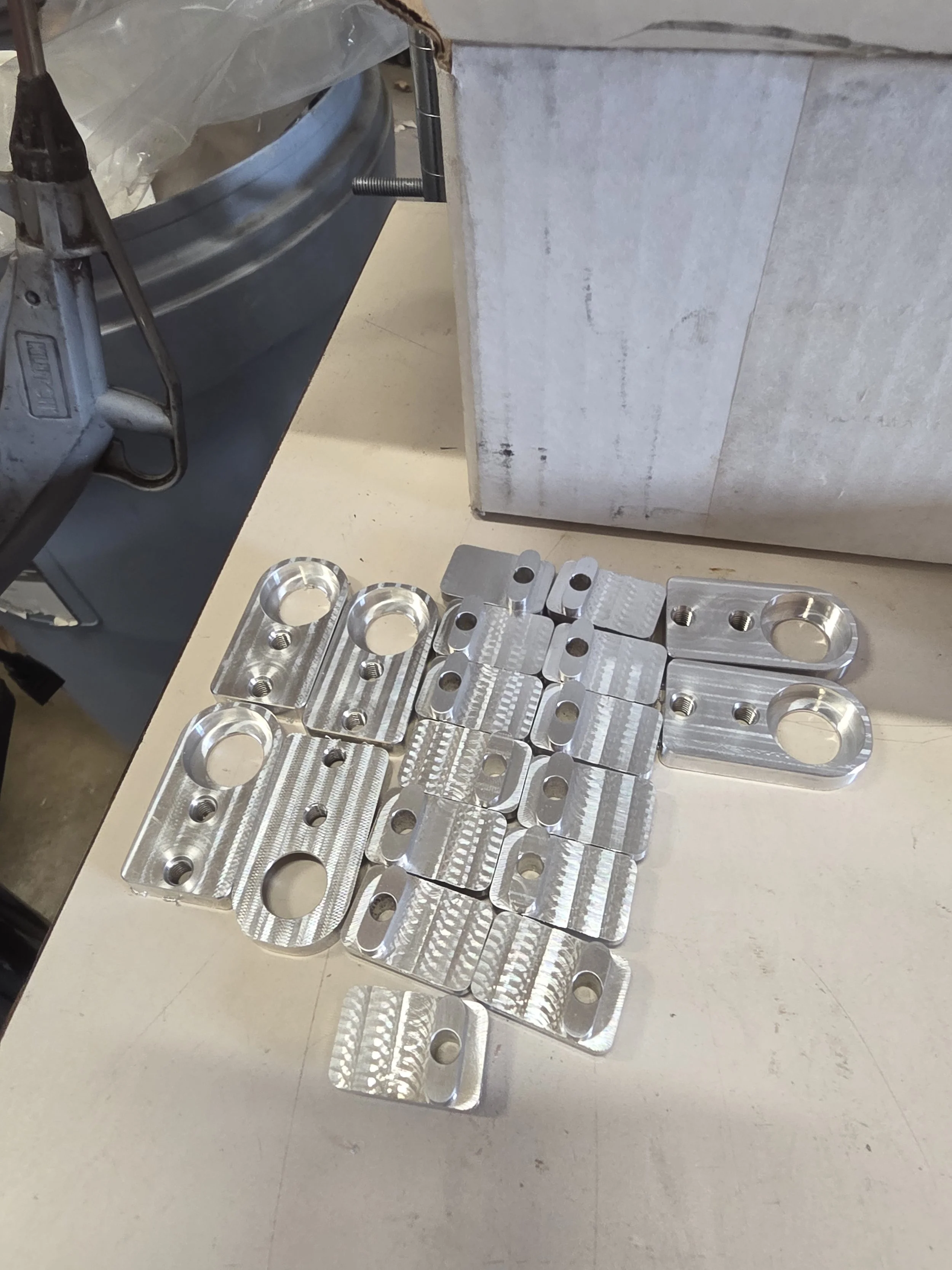 Multiple machined metal parts with holes and grooves laid out on a beige work surface.