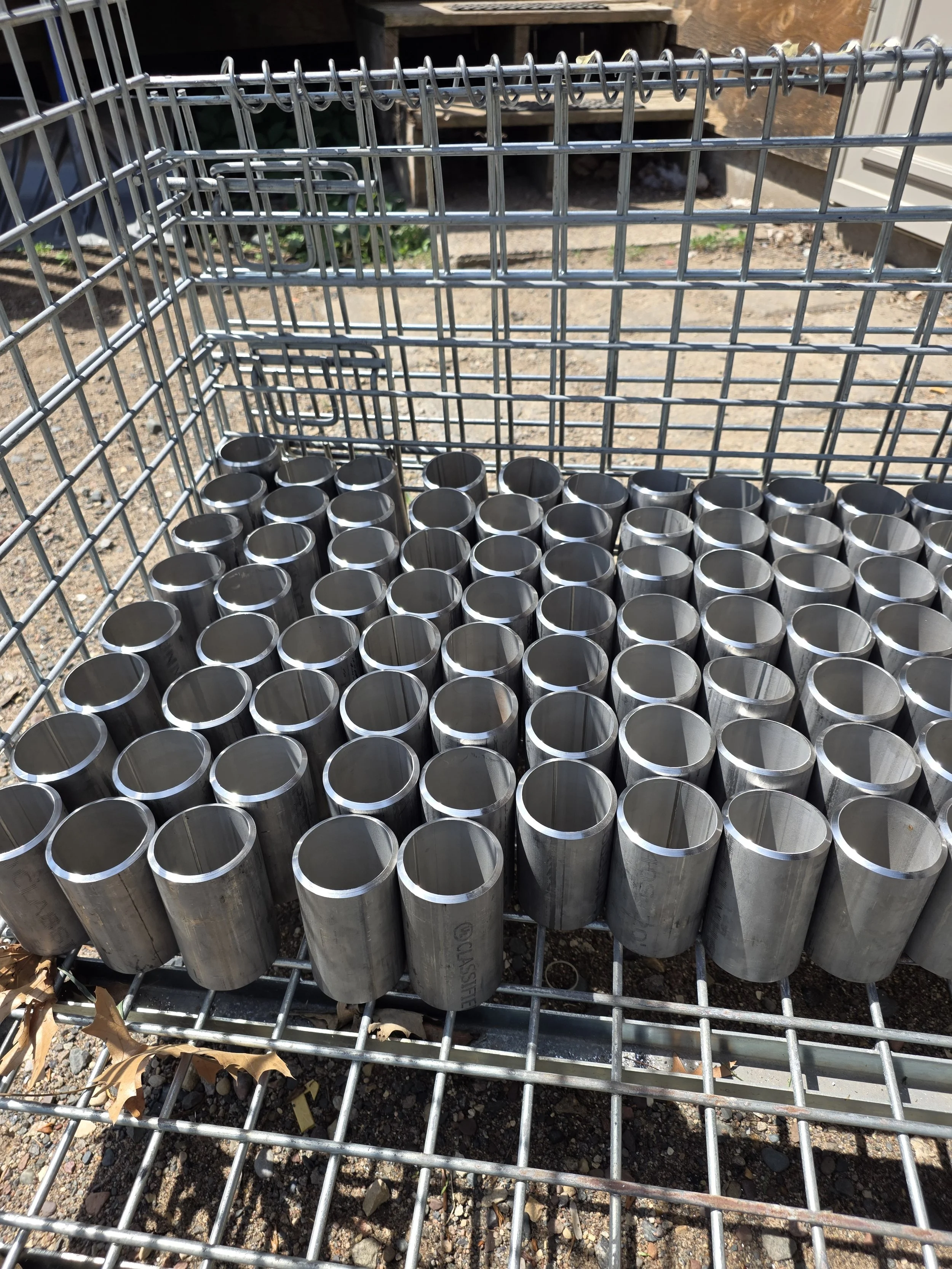 A wire cart holding multiple stainless steel cylinders outdoors on a gravel surface. Dry leaves are visible at the bottom of the image.