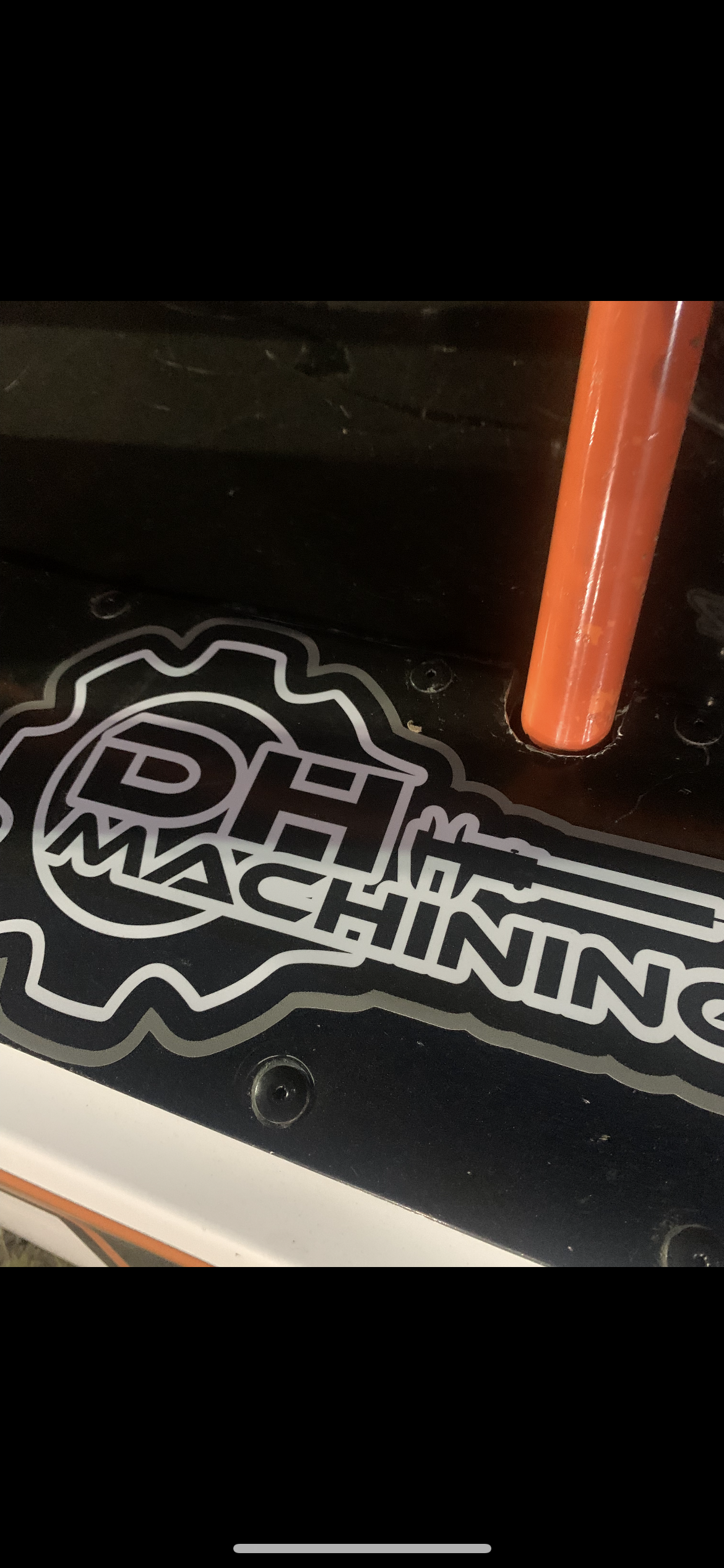 Close-up of a black surface with a sticker that says 'RACING' and 'MACHINING' and features a gear design, along with an orange metal rod attached to the surface.