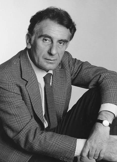 Black and white portrait of a man in a suit with a checked blazer, white shirt, and tie, sitting with one knee up and resting his arm on it, looking at the camera.