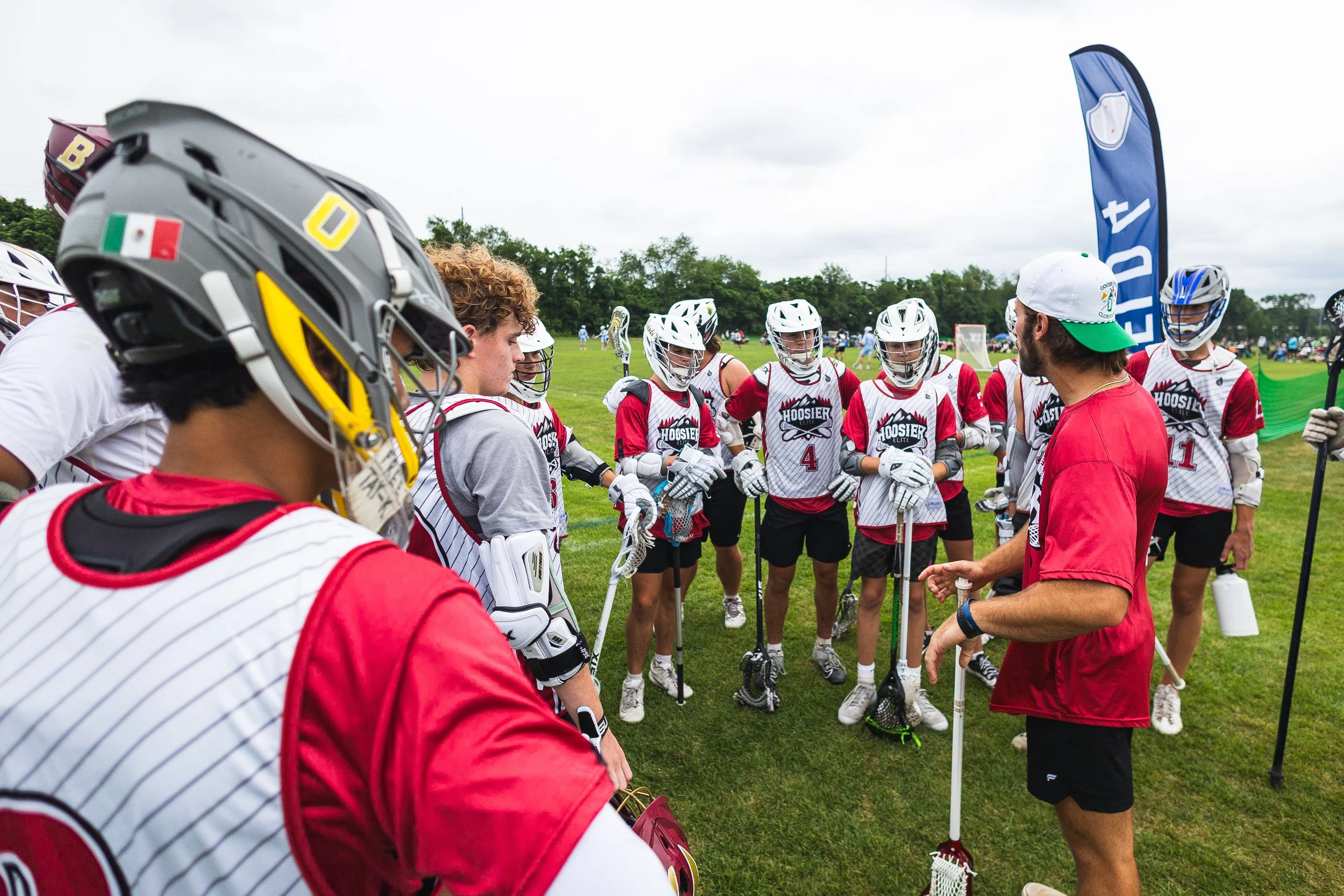 Lacrosse team huddling around their coach during practice on a grass field with overcast sky.
