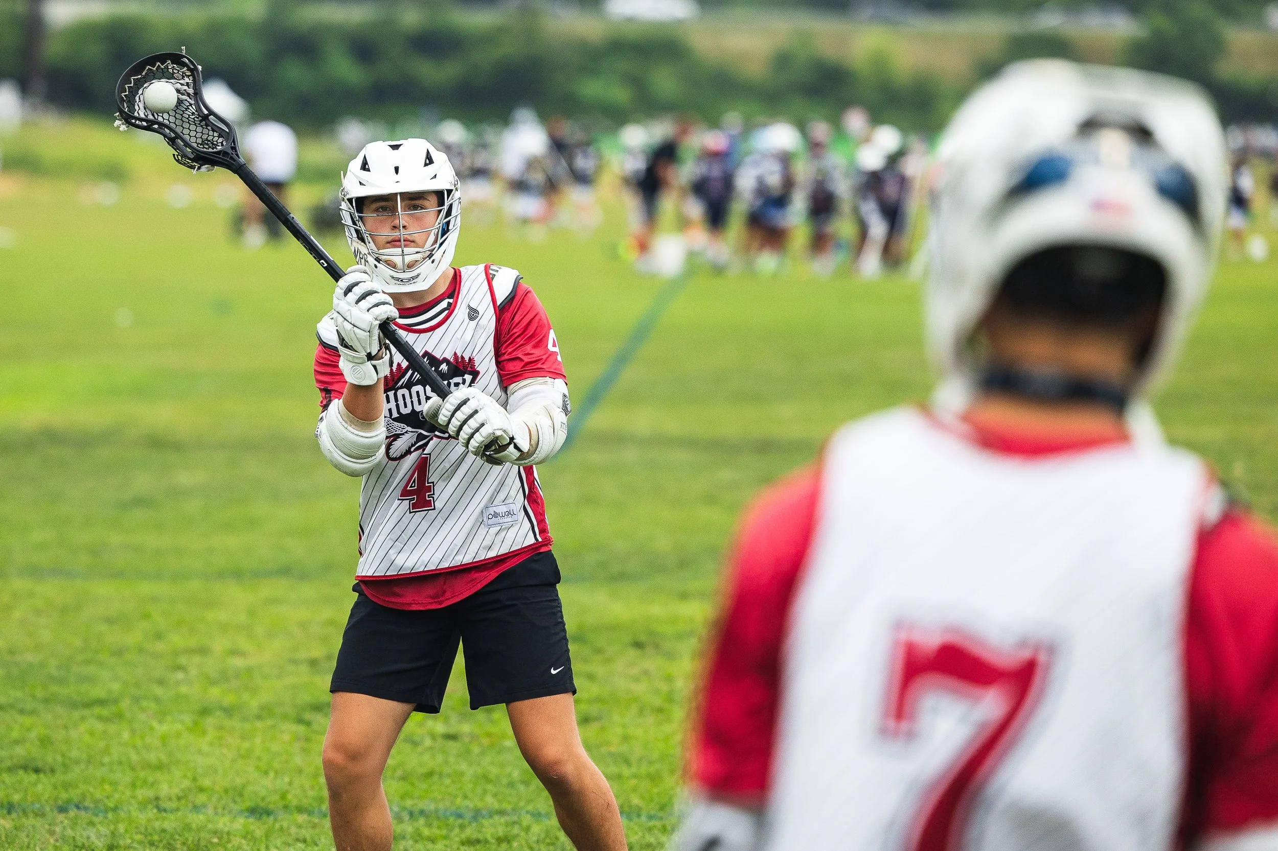 A lacrosse player wearing a white helmet, red and white jersey with the number 4, holding a lacrosse stick, stands on a grassy field during a game or practice.