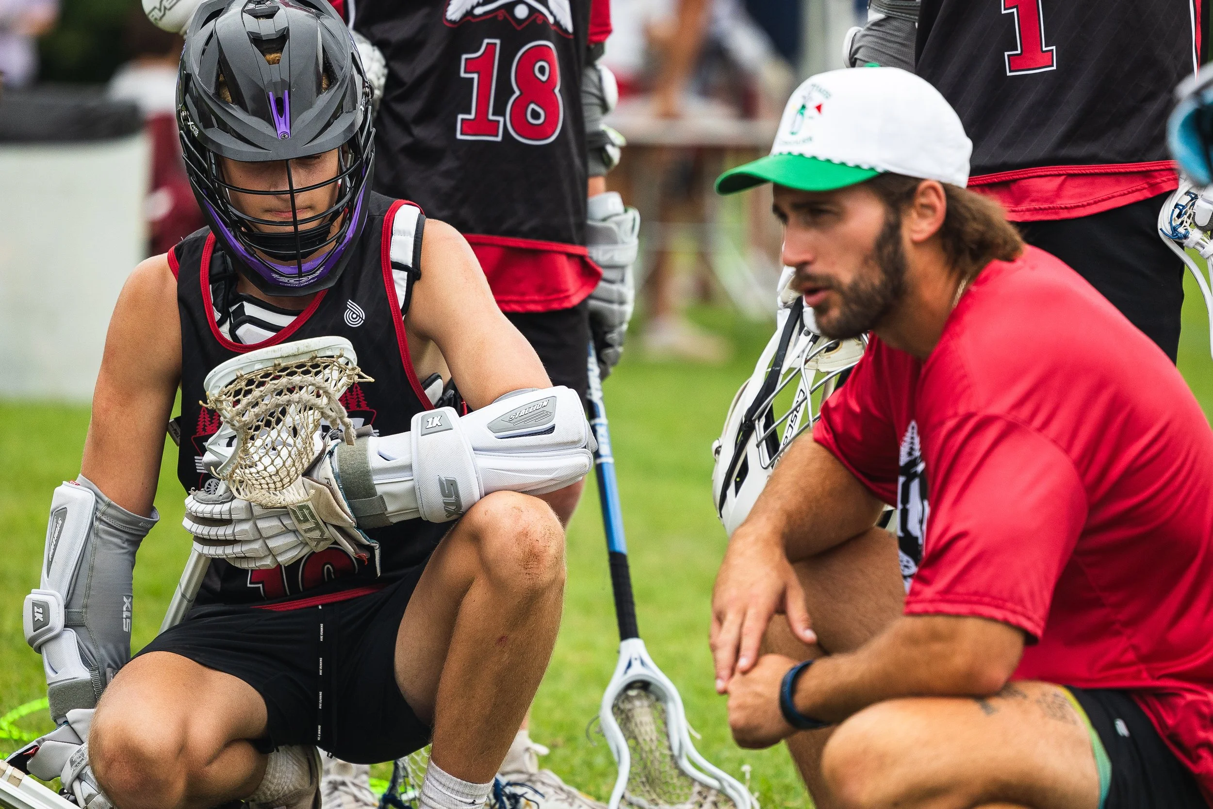 A lacrosse player with a helmet and gloves kneeling on the grass, listening to a coach giving instructions during a game or practice.