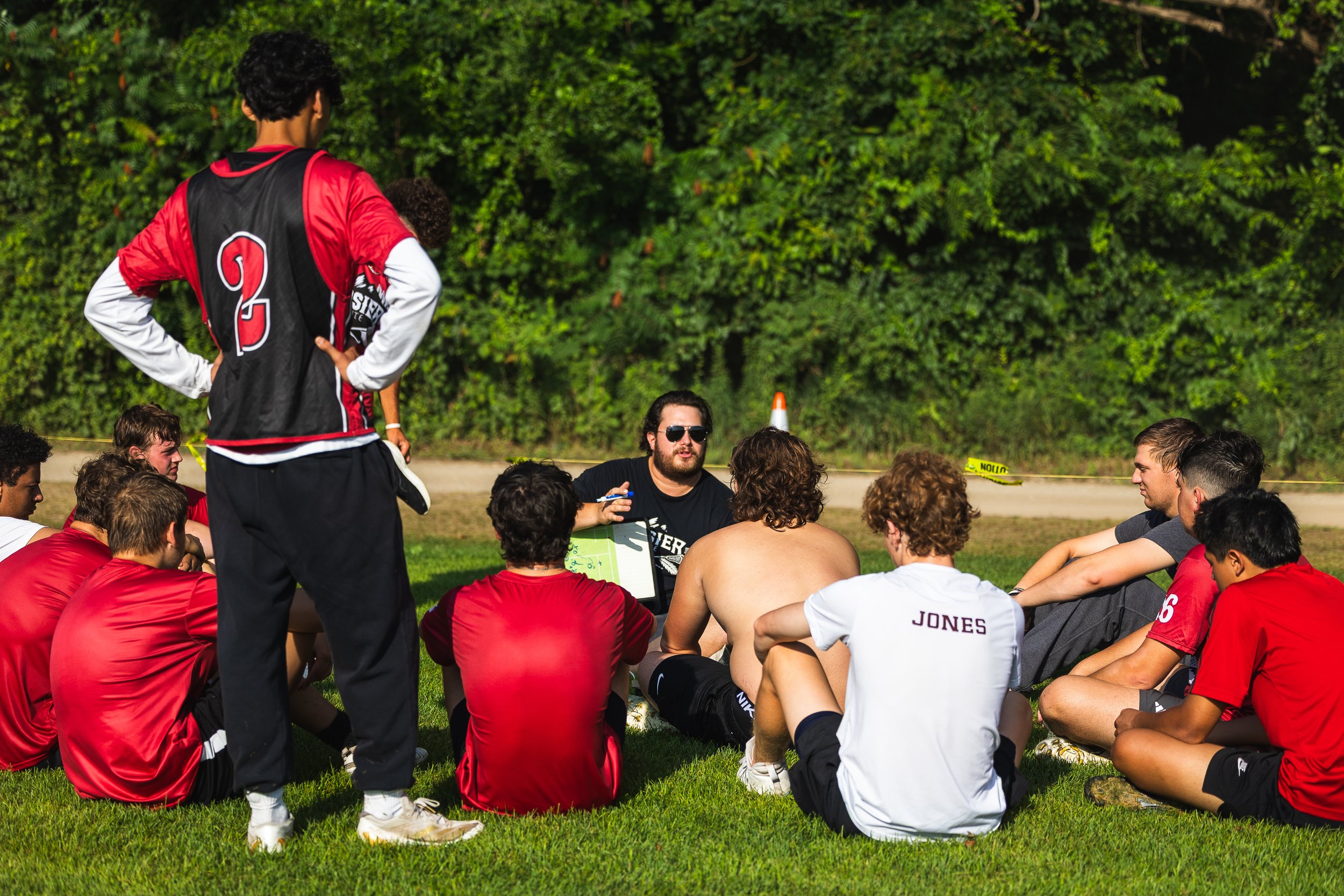 Group of young men, some shirtless, sitting on grass during a sports team meeting outdoors. One standing man, wearing a red and black sports jersey with the number 2, faces the group, while another man with sunglasses and a black shirt appears to be speaking or leading the discussion.