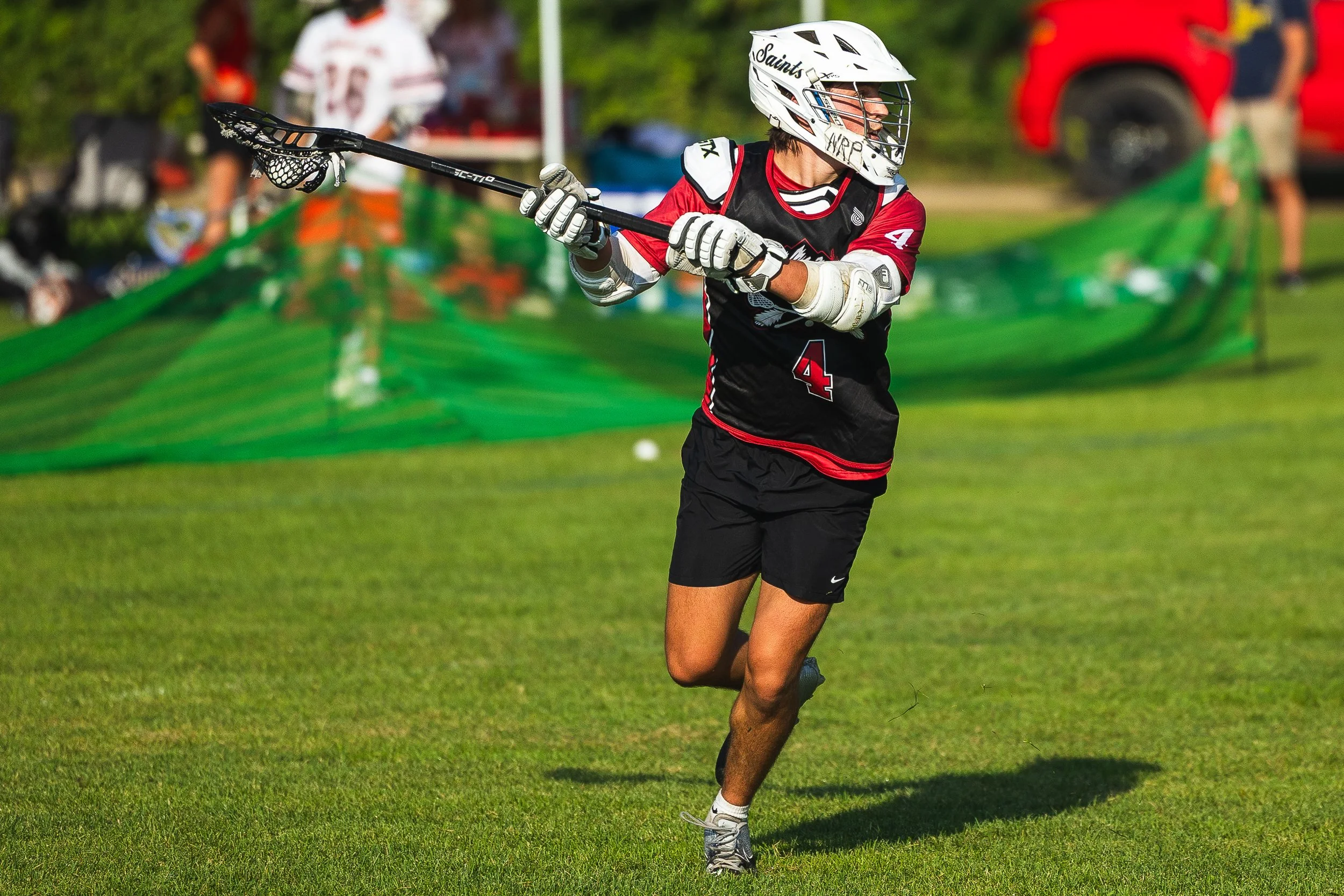 A lacrosse player in a black and red uniform, wearing a white helmet, running on a grassy field while holding a lacrosse stick.