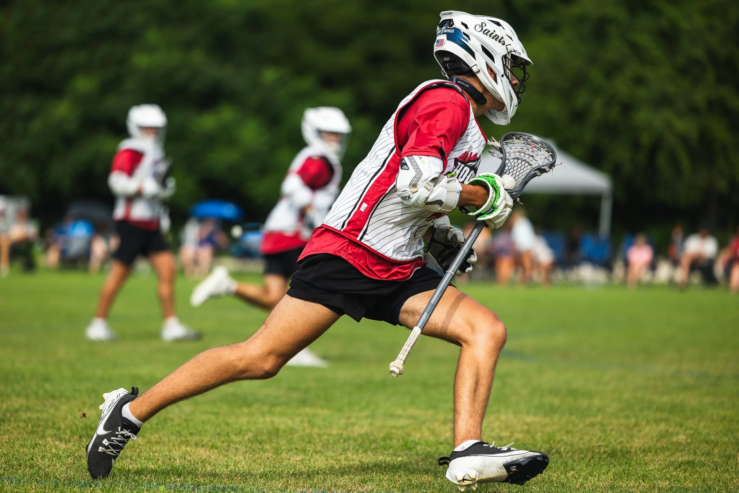 A lacrosse player running on a field with other players and spectators in the background, wearing a helmet, gloves, and athletic gear.