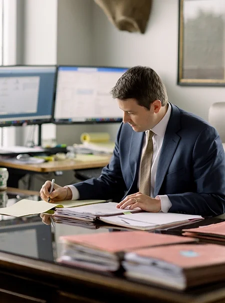 Businessman in a suit working at a cluttered desk with multiple documents, in an office with dual computer monitors.