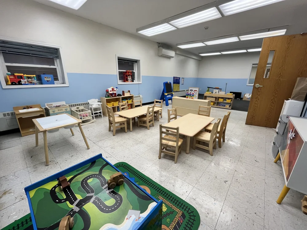 Tiny Tots classroom with child-sized wooden tables, shelves of books and toys, and a play mat with a road and train set