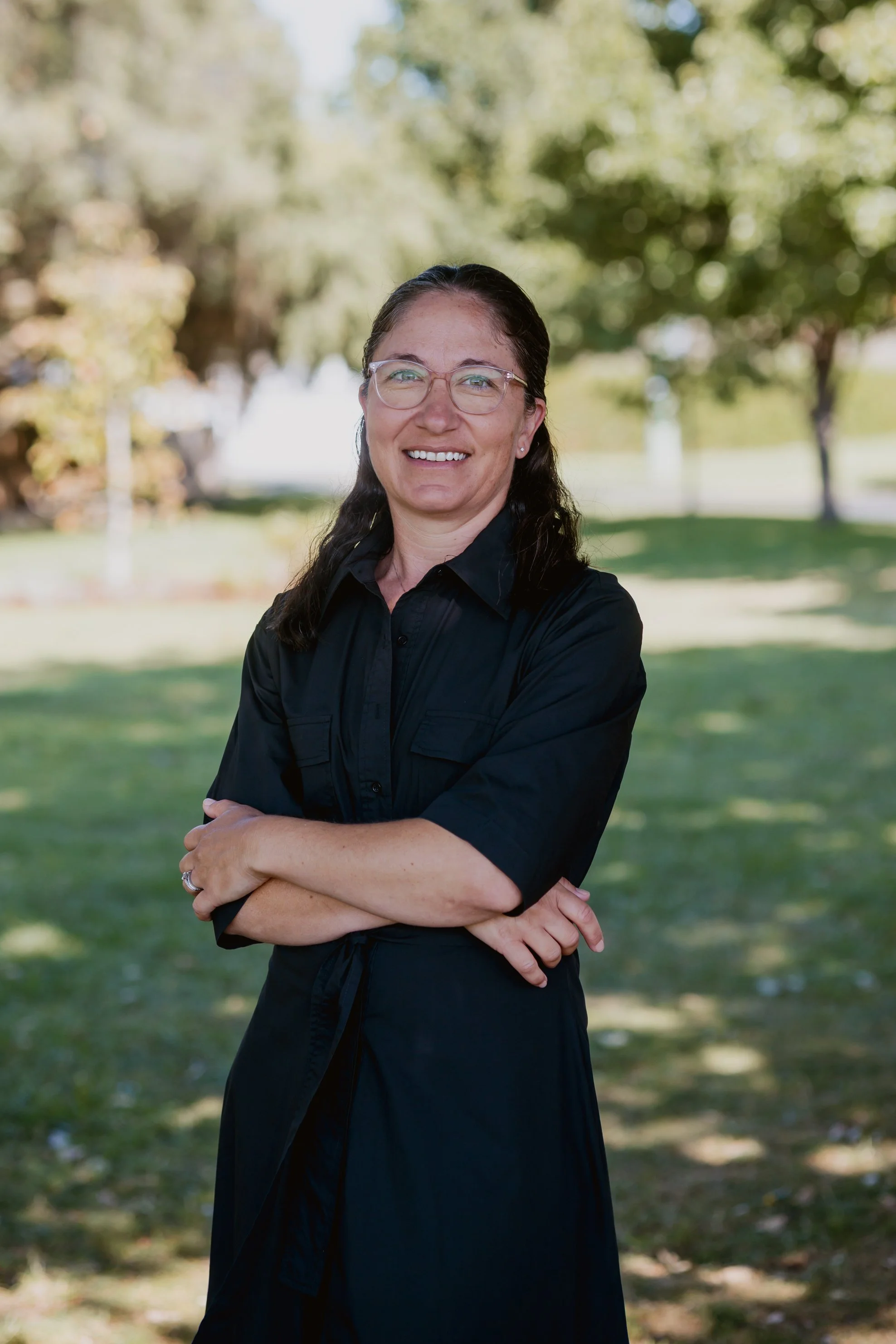 A woman with glasses and dark hair standing outdoors with arms crossed, smiling, in a park with trees and grass.