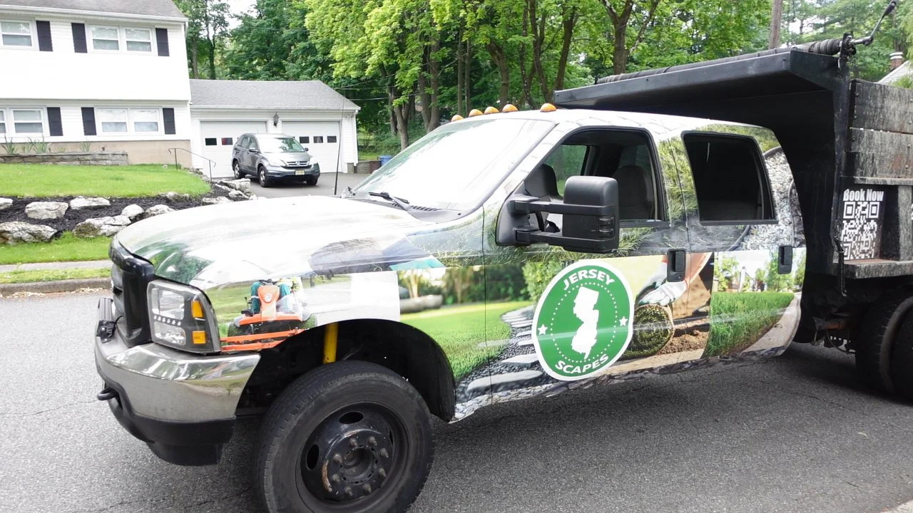 A truck with a Jersey Scapes logo parked on a residential street, with a house and another car in the background.