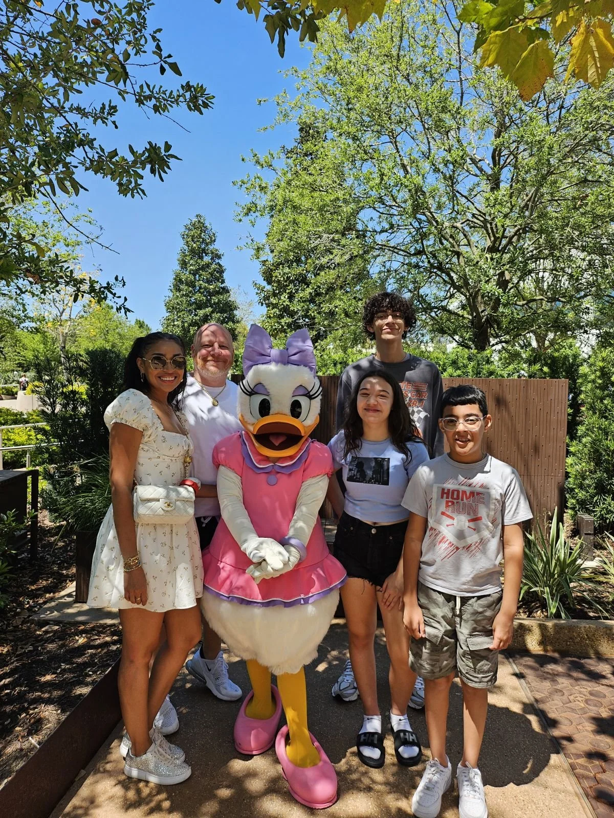 Group of six people and a person in a Donald Duck costume posing for photo outdoors on a sunny day, surrounded by trees and greenery.