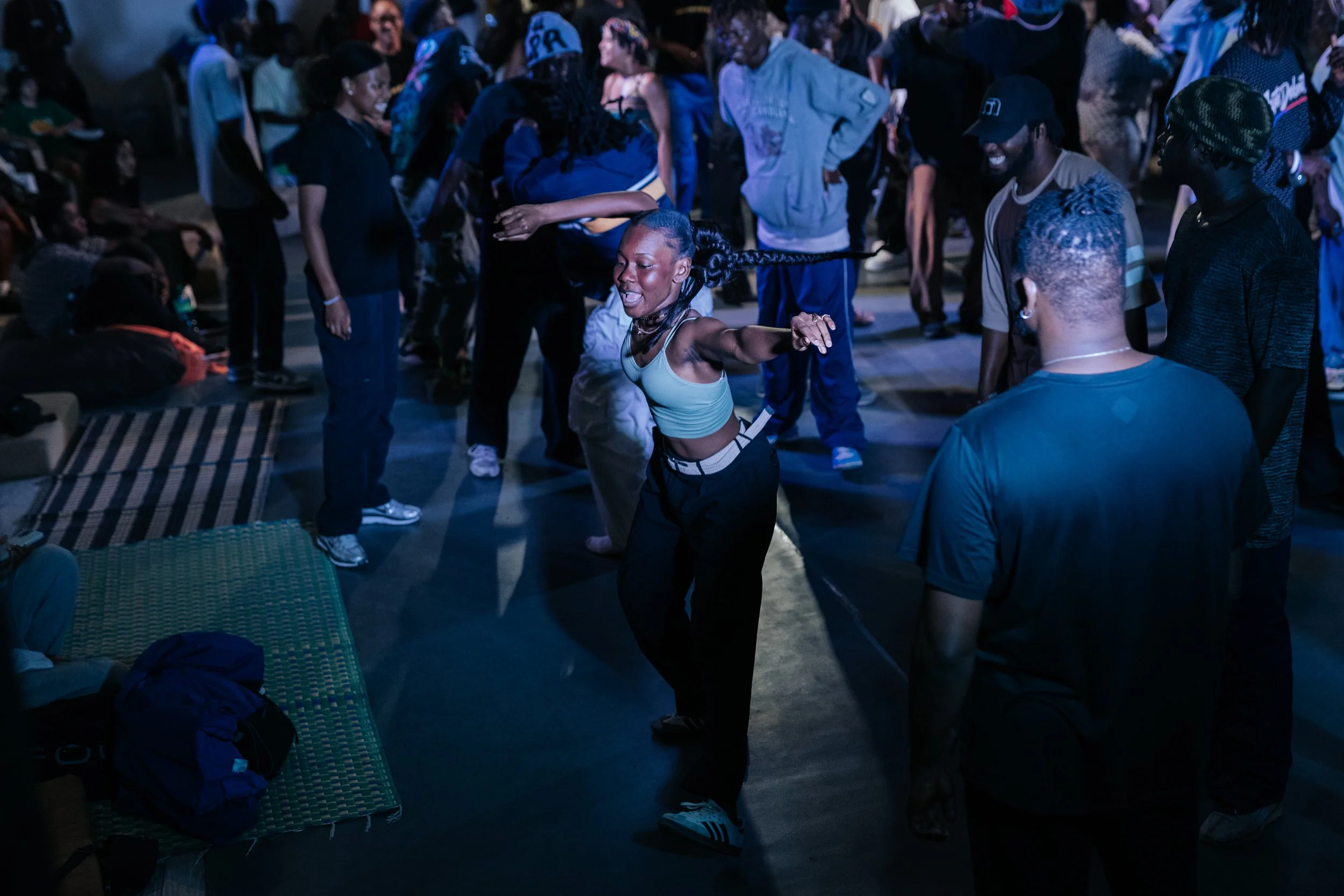 Une femme jeune danse au centre d'une foule en intérieur lors d'une soirée, entourée d'hommes et de femmes qui regardent et participent à la danse.