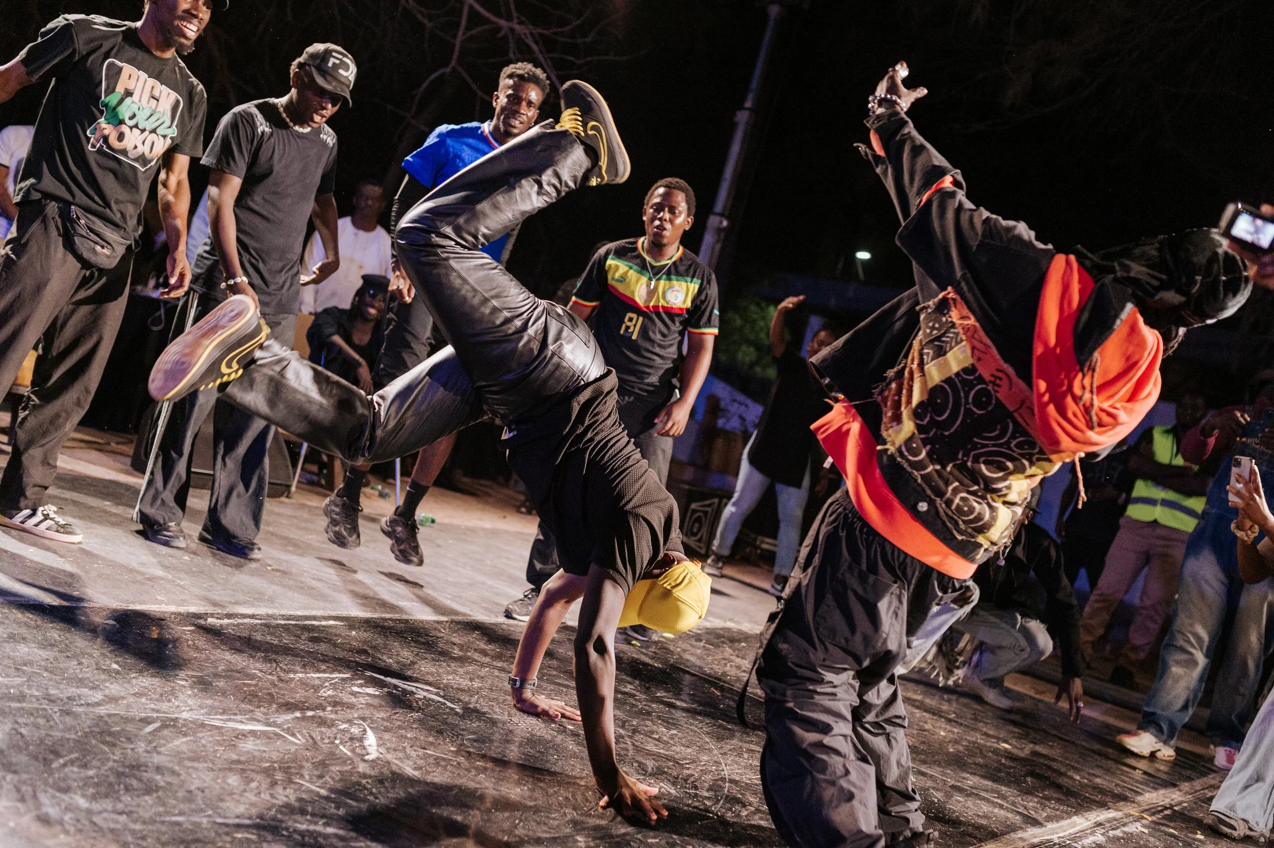 Des danseurs breakdance en pleine performance lors d'une soirée nocturne, avec un public qui les regarde et photographie.
