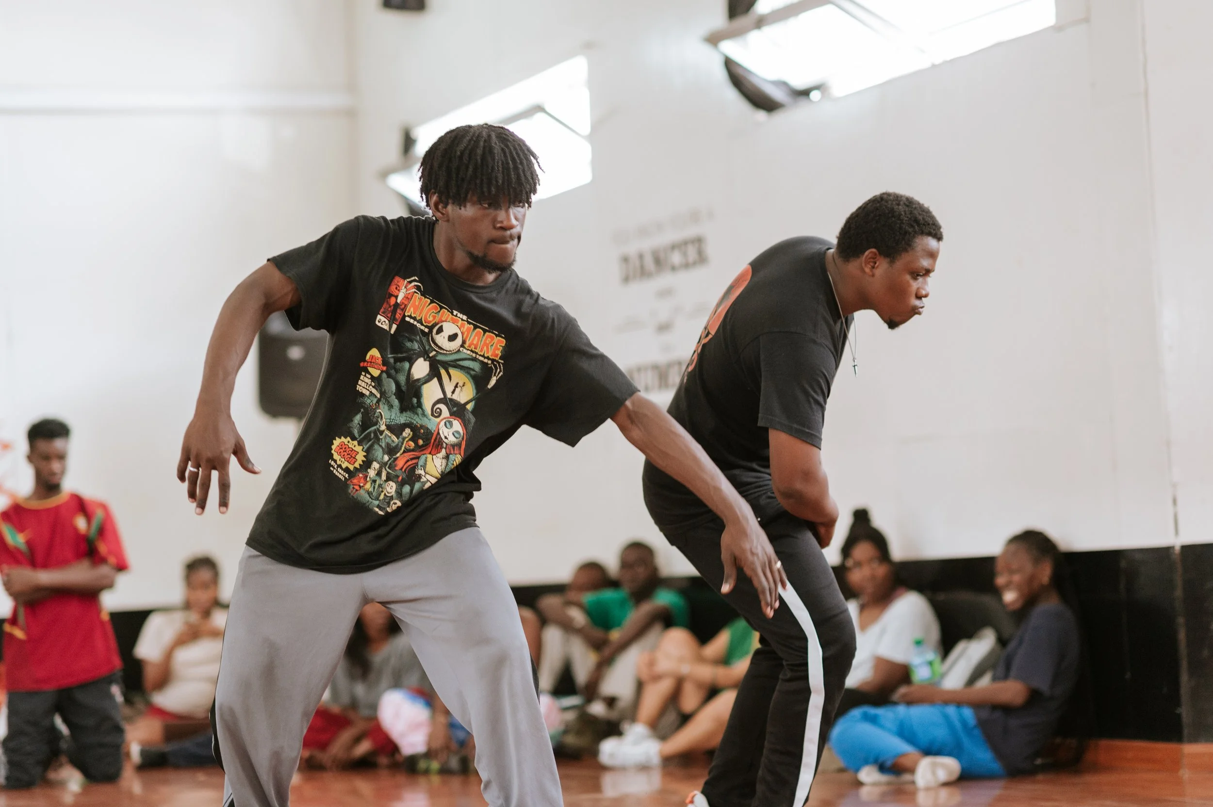 Deux jeunes hommes dansent dans une salle de danse, entourés de spectateurs assis en arrière-plan.