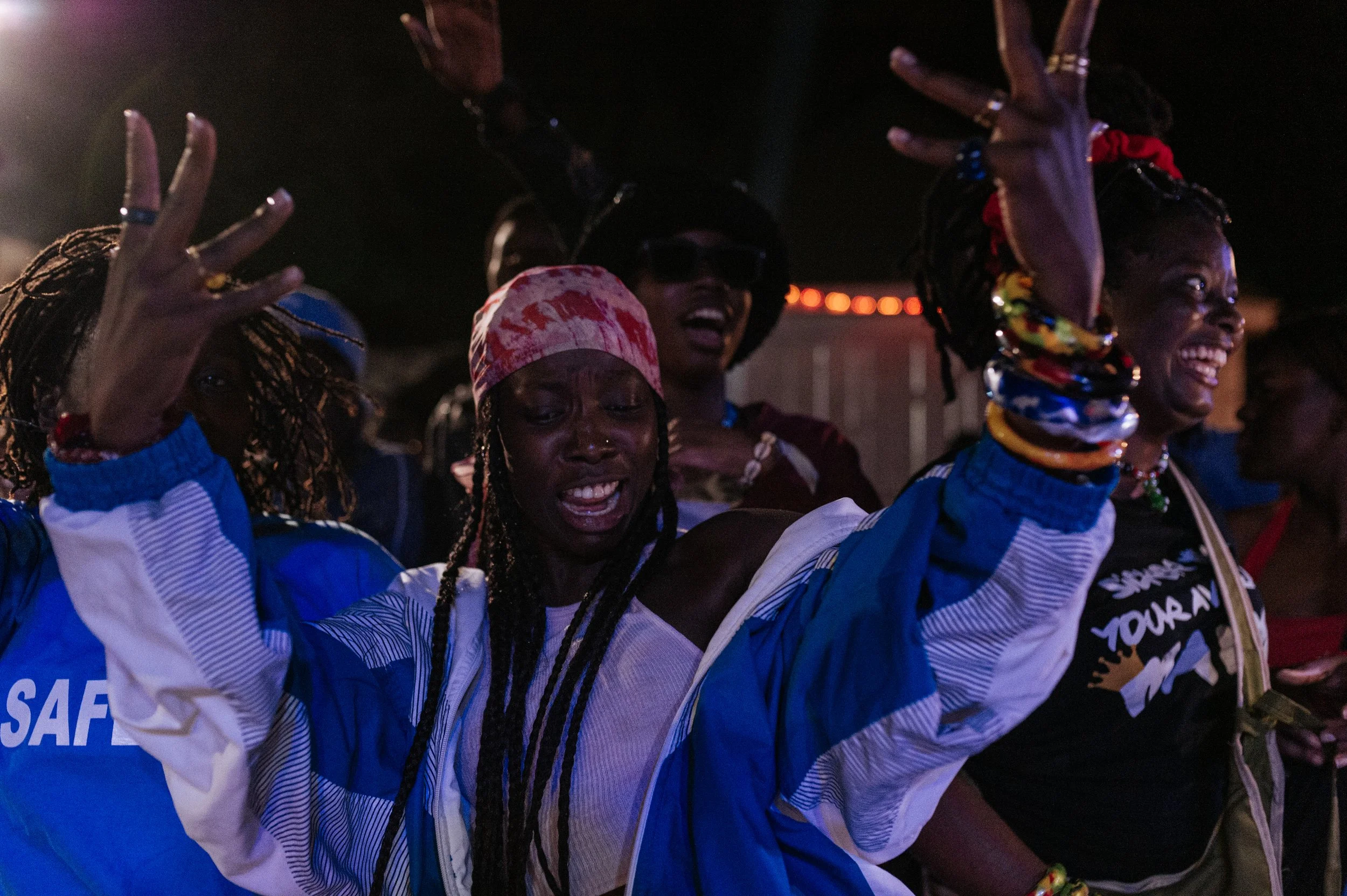 Groupe de femmes africaines souriantes en mouvement lors d'une célébration ou fête, portant des vêtements colorés et accessoires vibrants, dans une ambiance festive nocturne.