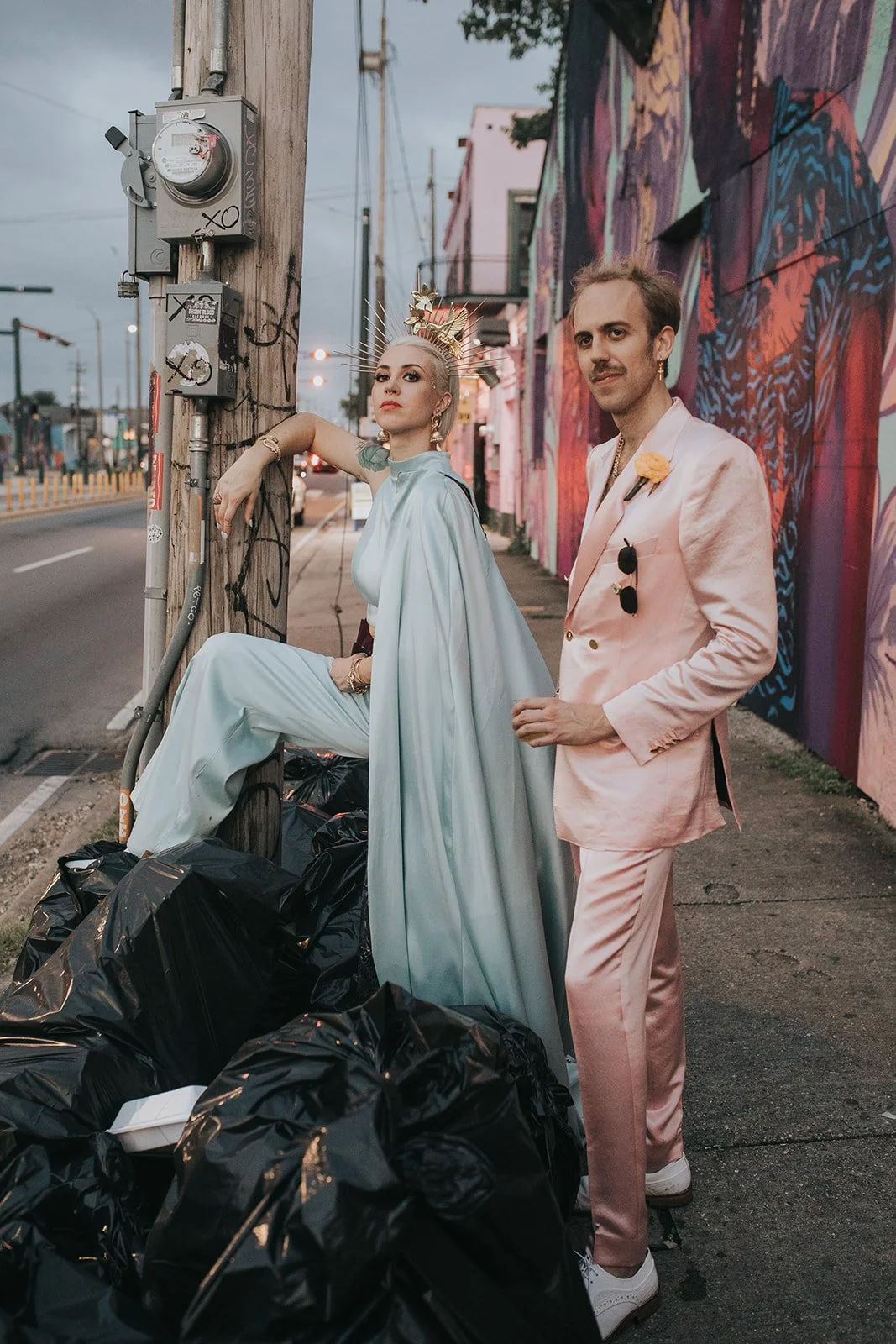 Baz is dressed in a blue 2 piece with a golden crown, and Matthew Wilder is in a pink suit with sunglasses and a flower boutonniere. They are standing on a New Orleans sidewalk next to a mural and a utility pole with trash bags at their feet.