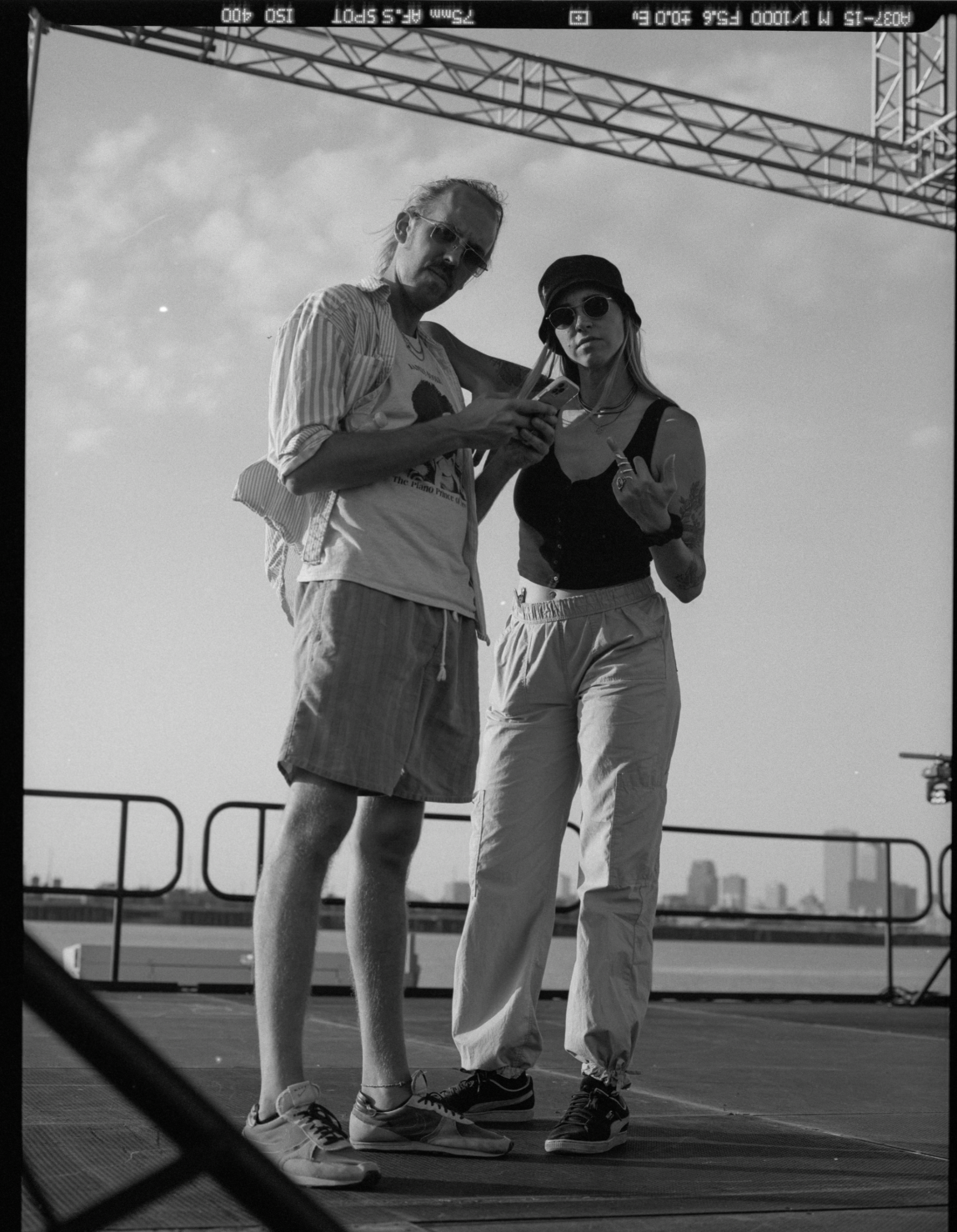 Baz & Wilder are standing outdoors on a rooftop, with a city skyline in the background. They are wearing casual summer clothing and sunglasses, and are looking down at a smartphone held by the man. The woman is making a hand gesture.
