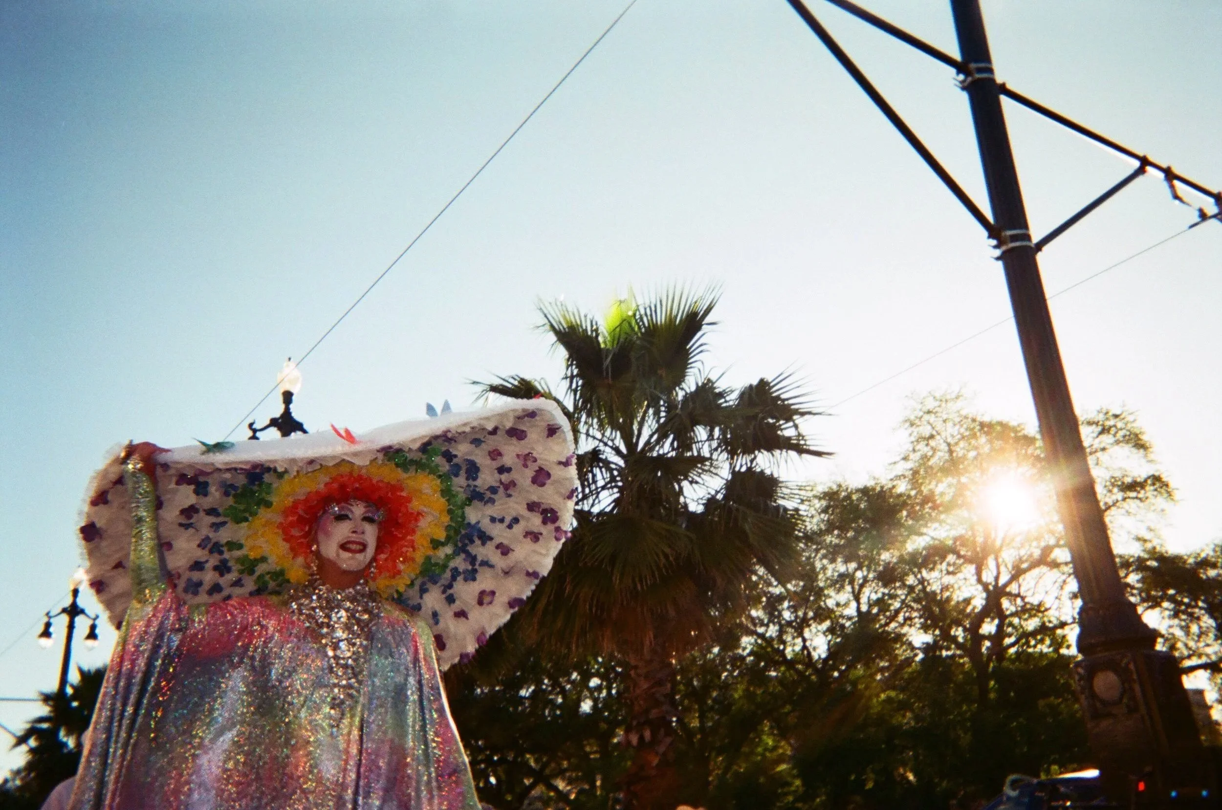 A performer at the New Orleans Gay Easter parade is dressed in a colorful, shiny costume with a large decorated headpiece, smiling while walking outdoors during daylight with trees and power lines in the background. Photo taken by Amber "Baz" Vogts