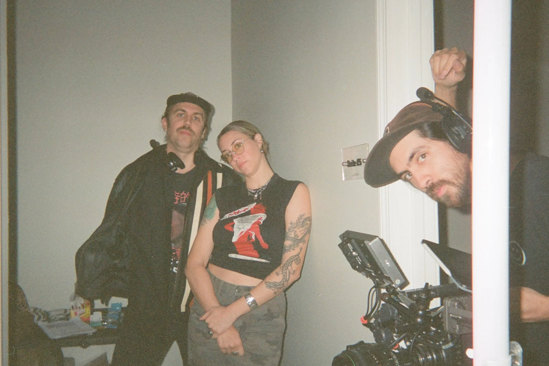 Baz & Wilder are stood in a small room, along with a Director of Photography.  The Director of Photography has long dark hair, a beard, and is wearing a hat, looking into the camera.