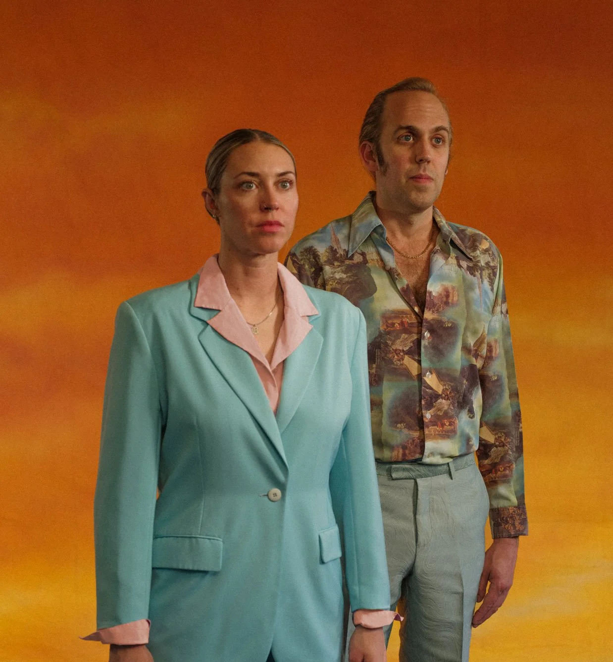 A woman, Baz, in a teal suit and a man, Matthew Wilder, in a patterned shirt stand side by side against a sunset backdrop background, both looking serious.