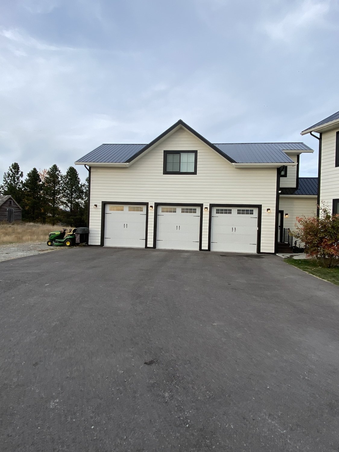White three-car garage with black accents and a gray roof, situated in a rural area with trees and an open field nearby.
