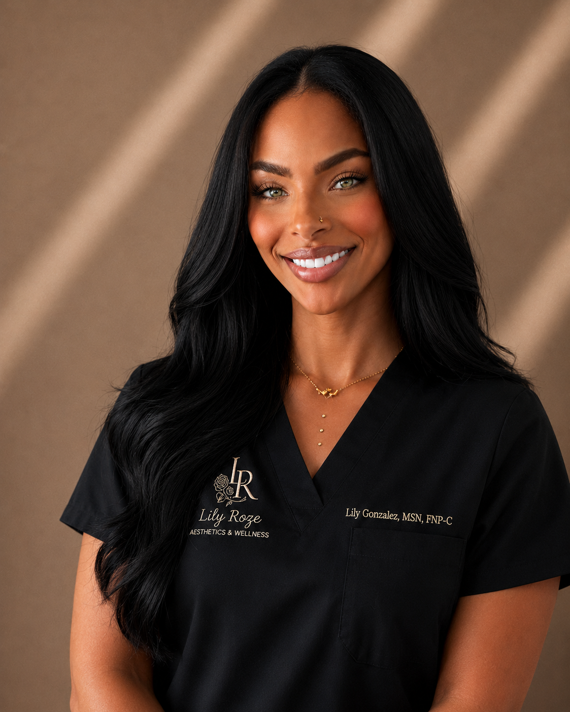 A woman with long black hair smiling, wearing a black medical scrub with embroidered name and title, gold jewelry, and a tan background.