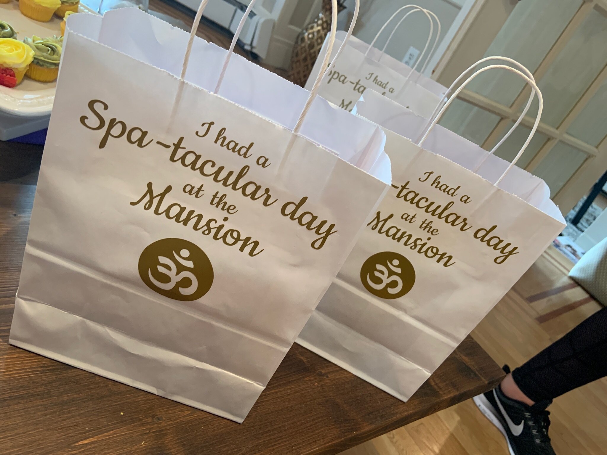 White gift bags with the text 'I had a Spa-tacular day at the Mansion' and an Om symbol, placed on a wooden table next to a plate of cupcakes.