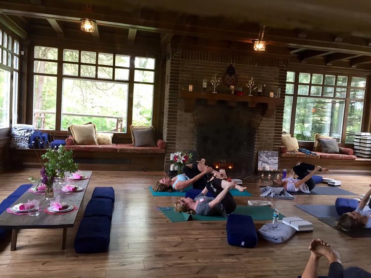 People practicing yoga in a cozy room with a fireplace, wooden floor, large windows, and a table set for a meal.