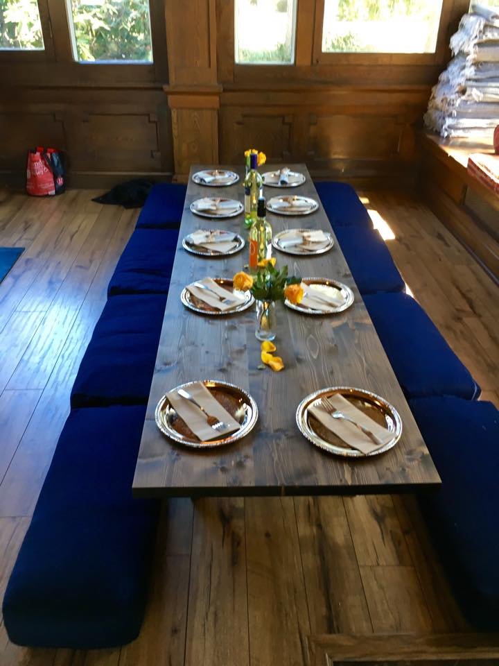 A low wooden dining table set for a meal, surrounded by blue floor cushions. The table is arranged with plates, napkins, and silverware. Three vases with yellow flowers and two bottles of wine are placed along the center. The setting is in a wooden r