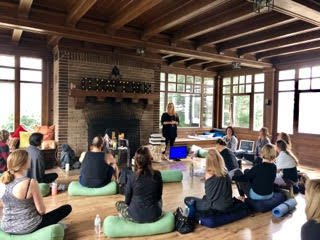 A group of people attending a workshop or class in a wooden room with large windows. They are seated on mats and pillows on the floor, facing a standing instructor who is giving a presentation. The room features a large brick fireplace and has a cozy