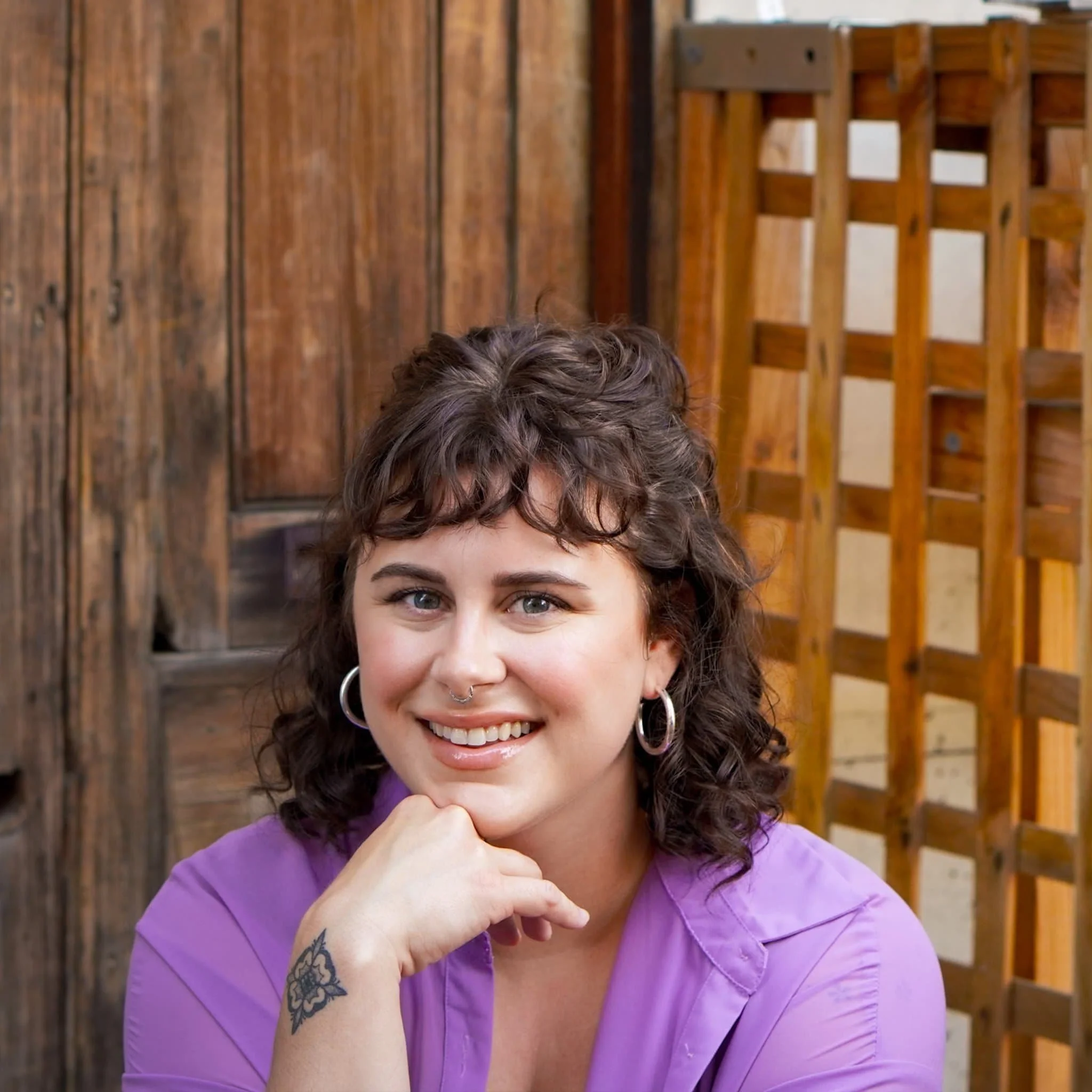 A smiling LGBTQ therapist with curly brown hair and hoop earrings sits by a wooden wall, wearing a purple shirt and resting her chin.