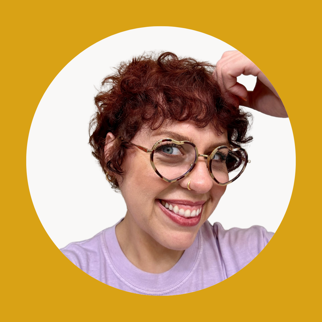 A woman with curly red hair, large circular glasses, a septum piercing, and a purple shirt, smiling and touching her hair against a white background with a mustard-colored border.