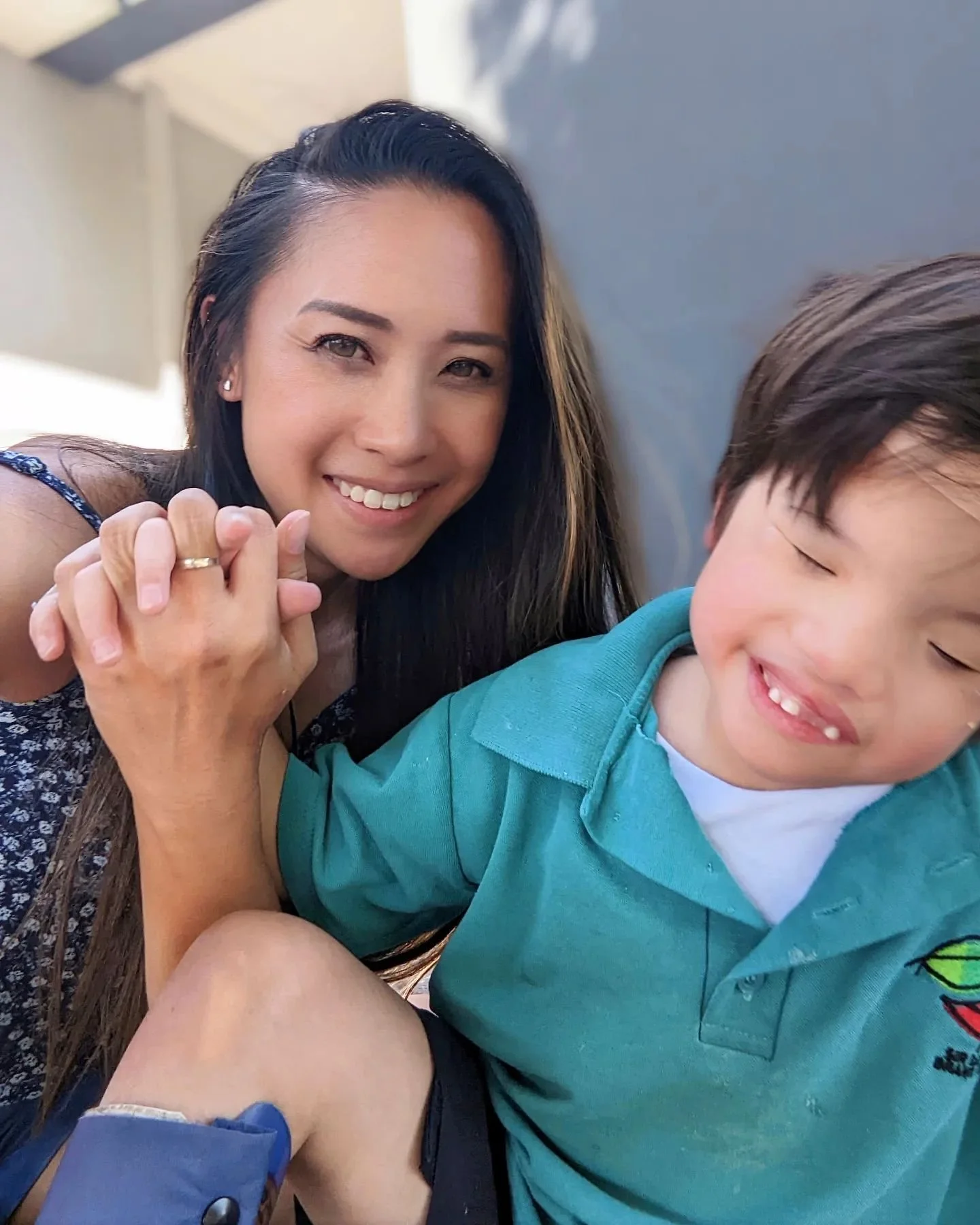A woman with long dark hair smiling and holding hands with a young boy with closed eyes and smiling, wearing a teal shirt, outdoors near a wall.
