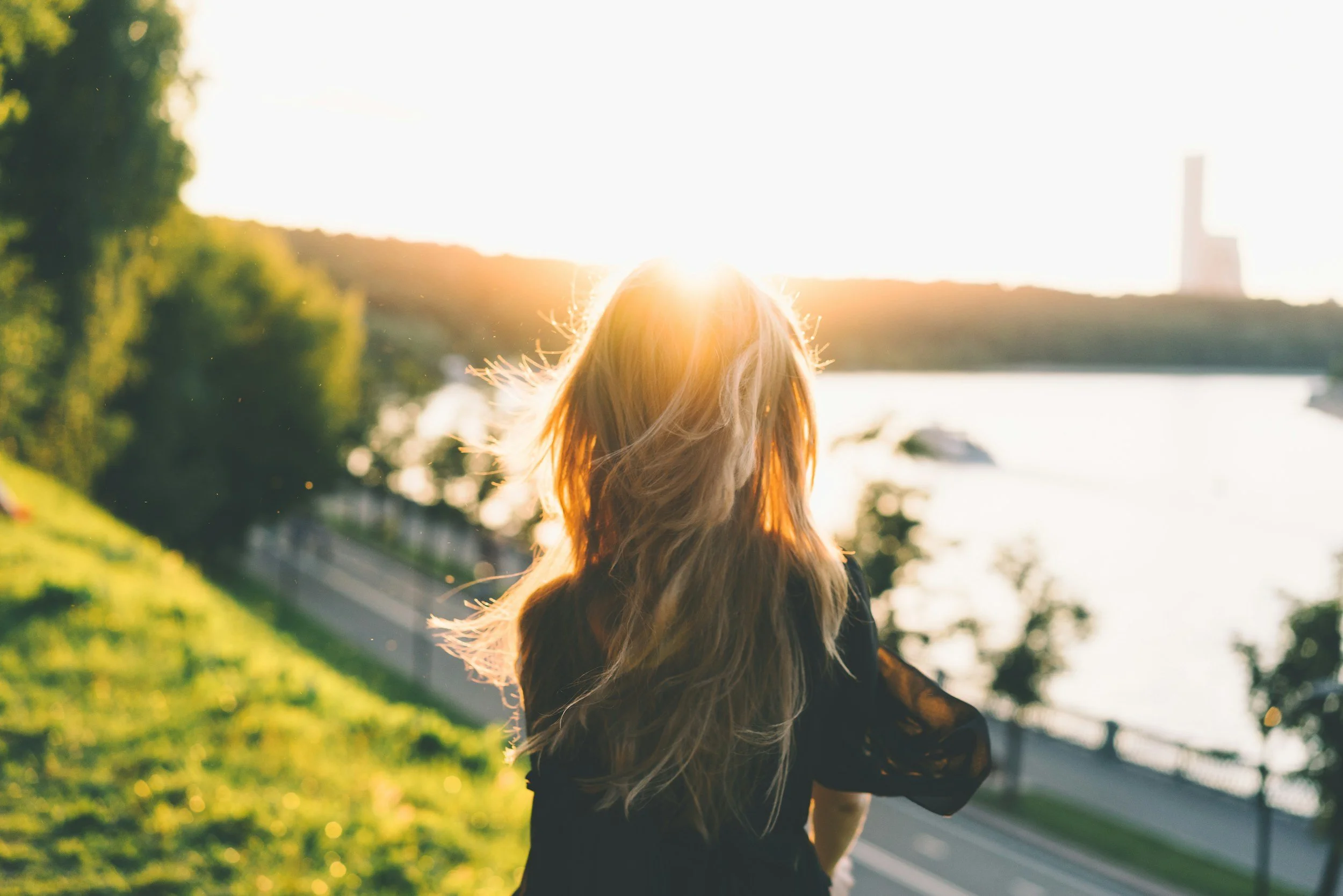 A woman with long, wavy hair standing outdoors at sunset near a river, looking away from the camera and facing the horizon.