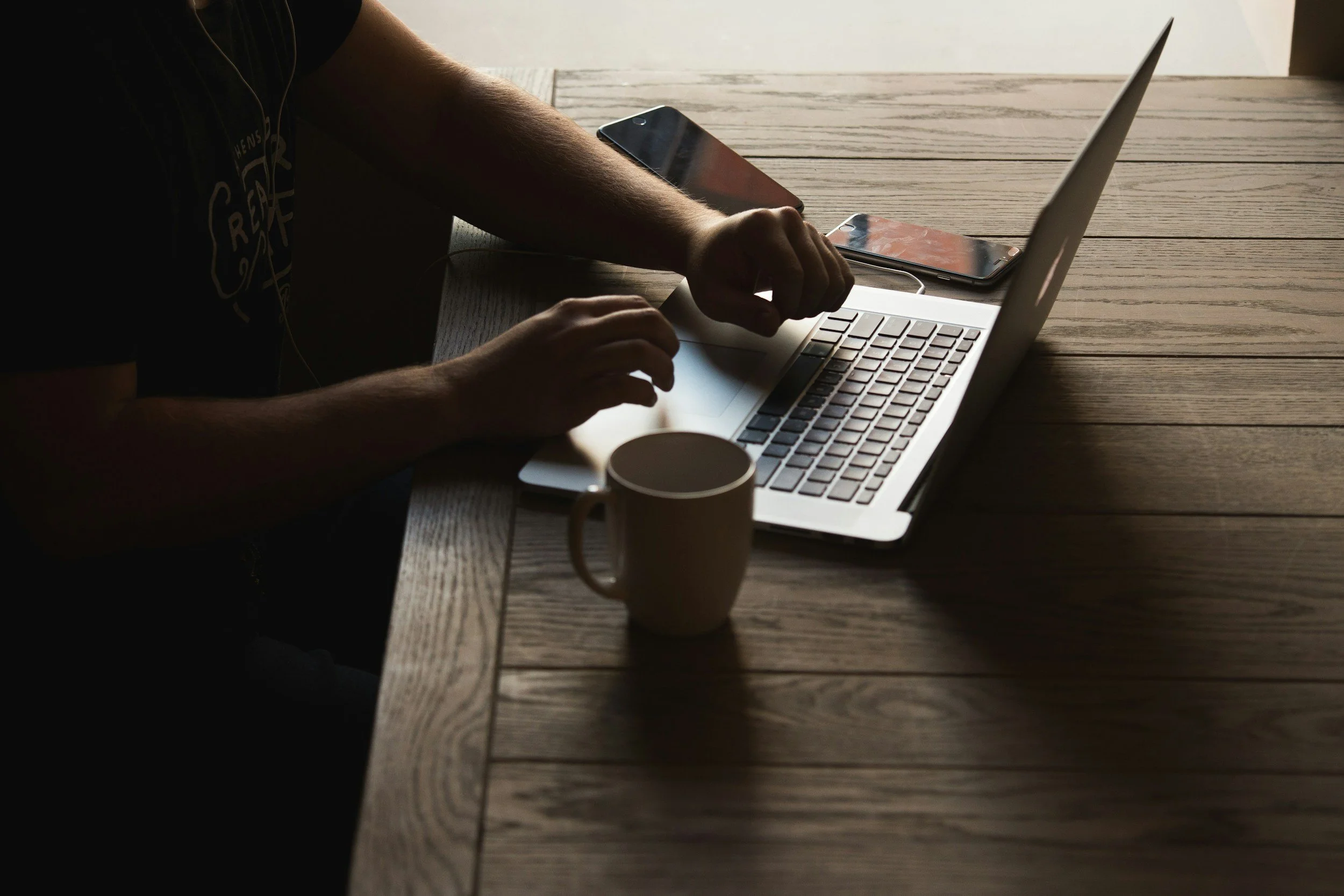A person working on a silver laptop at a wooden table, with a coffee mug and two smartphones nearby.