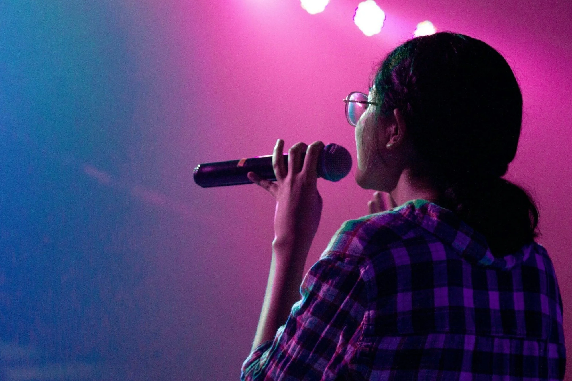 A woman with glasses holding a microphone and singing on stage with pink and blue lighting.