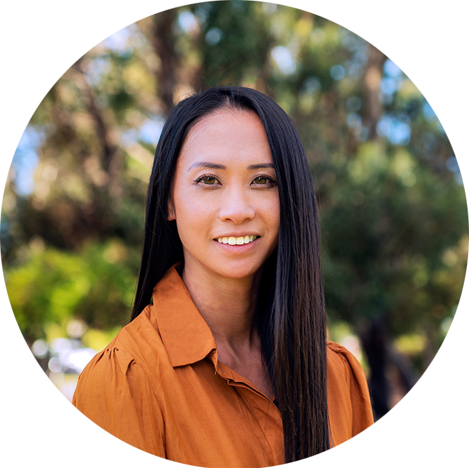 A smiling woman with long black hair wearing an orange blouse outdoors with blurred green trees in the background.