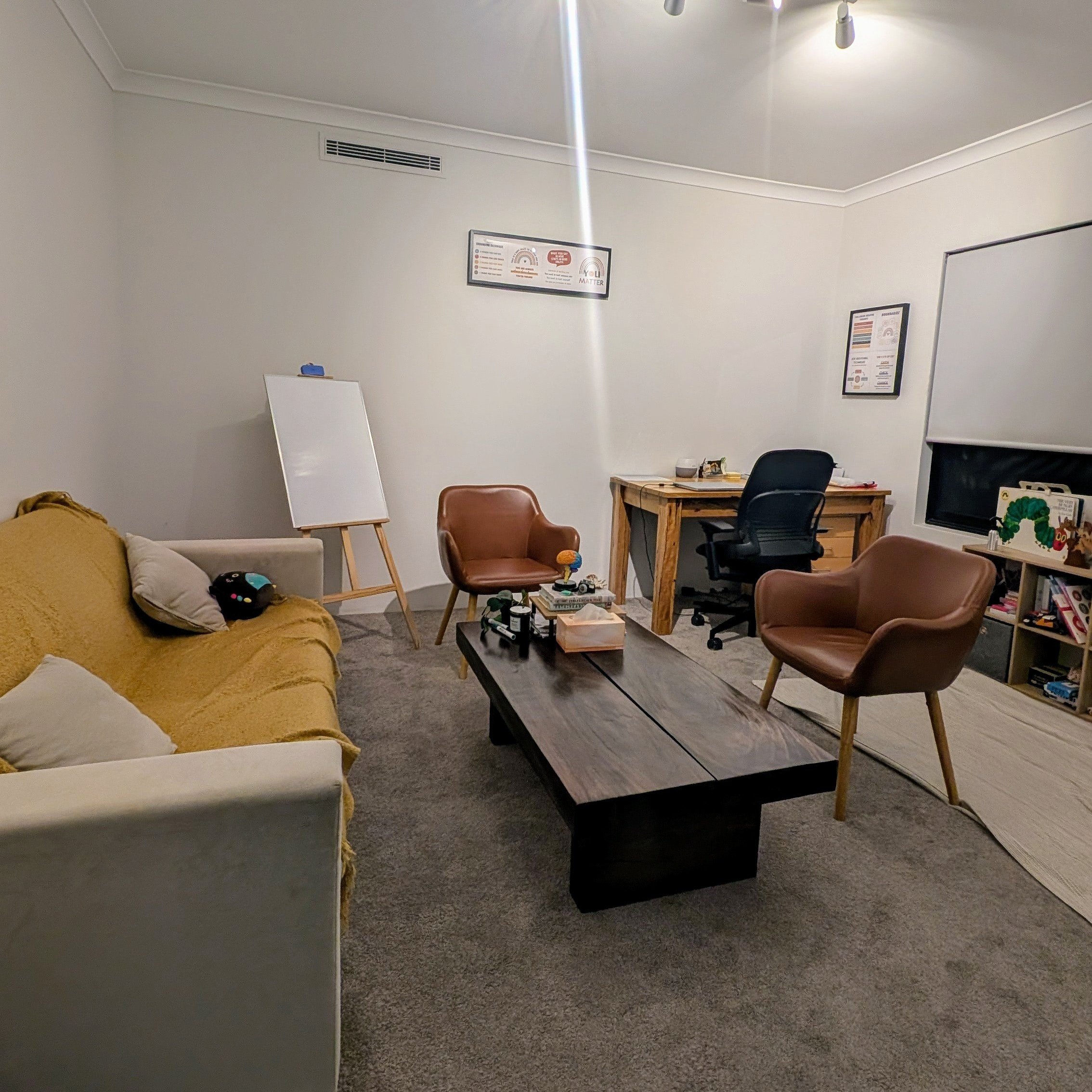 A cozy office or living room with a sofa, two brown armchairs, a black coffee table, a wooden desk with a black office chair, and a whiteboard on a stand. The room has beige carpet, white walls, and framed posters. There are toys and decorations on shelves, and a window with blinds.