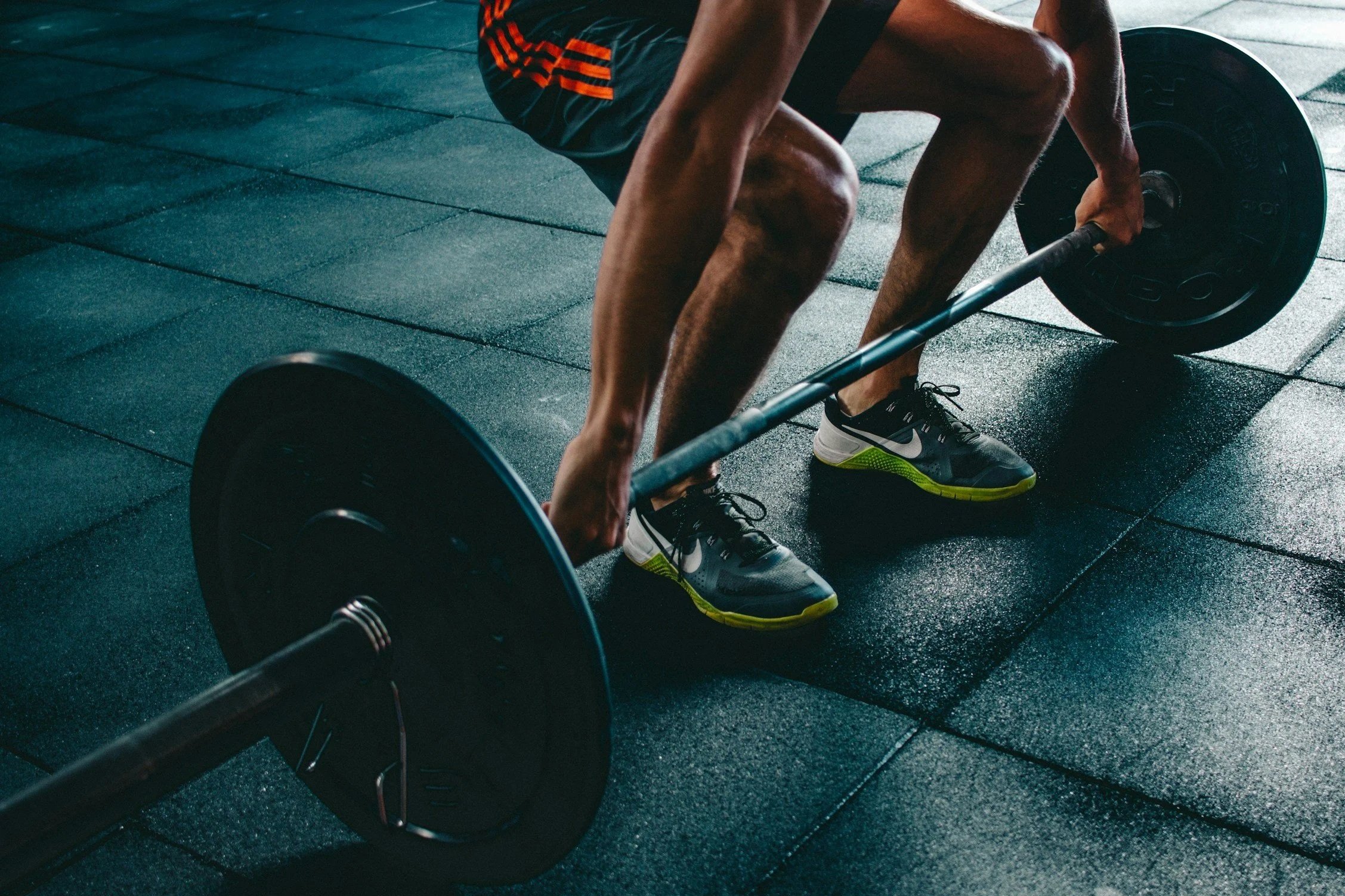 Person lifting a barbell with weights on a gym floor, wearing black Nike sneakers with neon green soles.