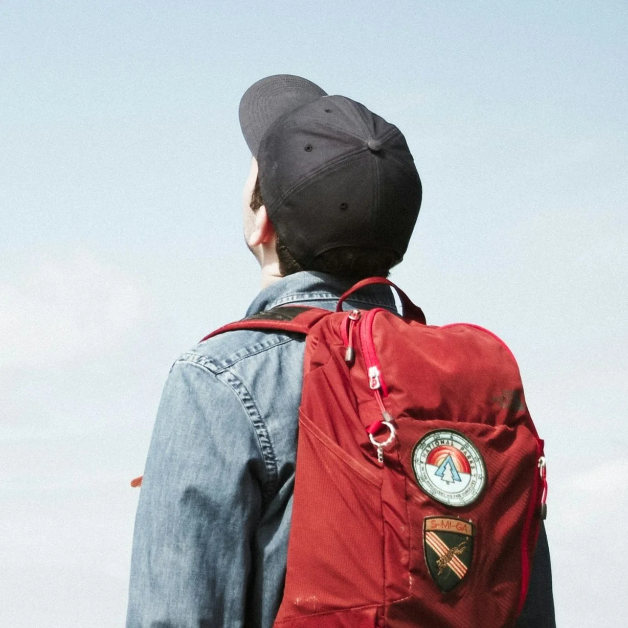 Person wearing a black cap and denim jacket with a red backpack, facing away against a cloudy sky.
